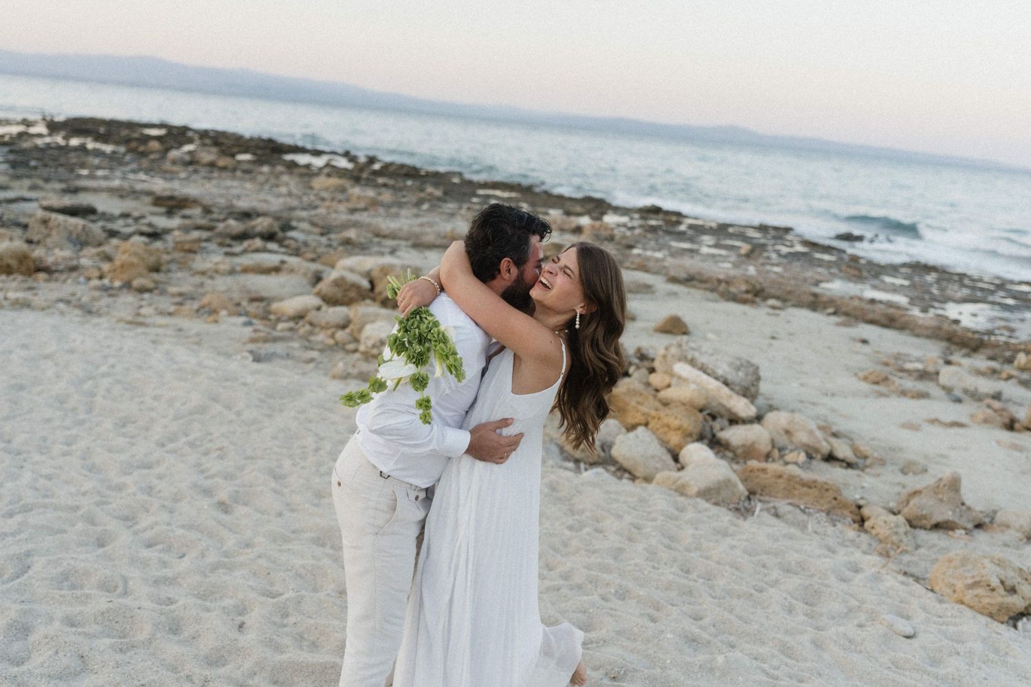 Couple in white embracing and kissing on a sandy beach with rocky shoreline and ocean in the background.
