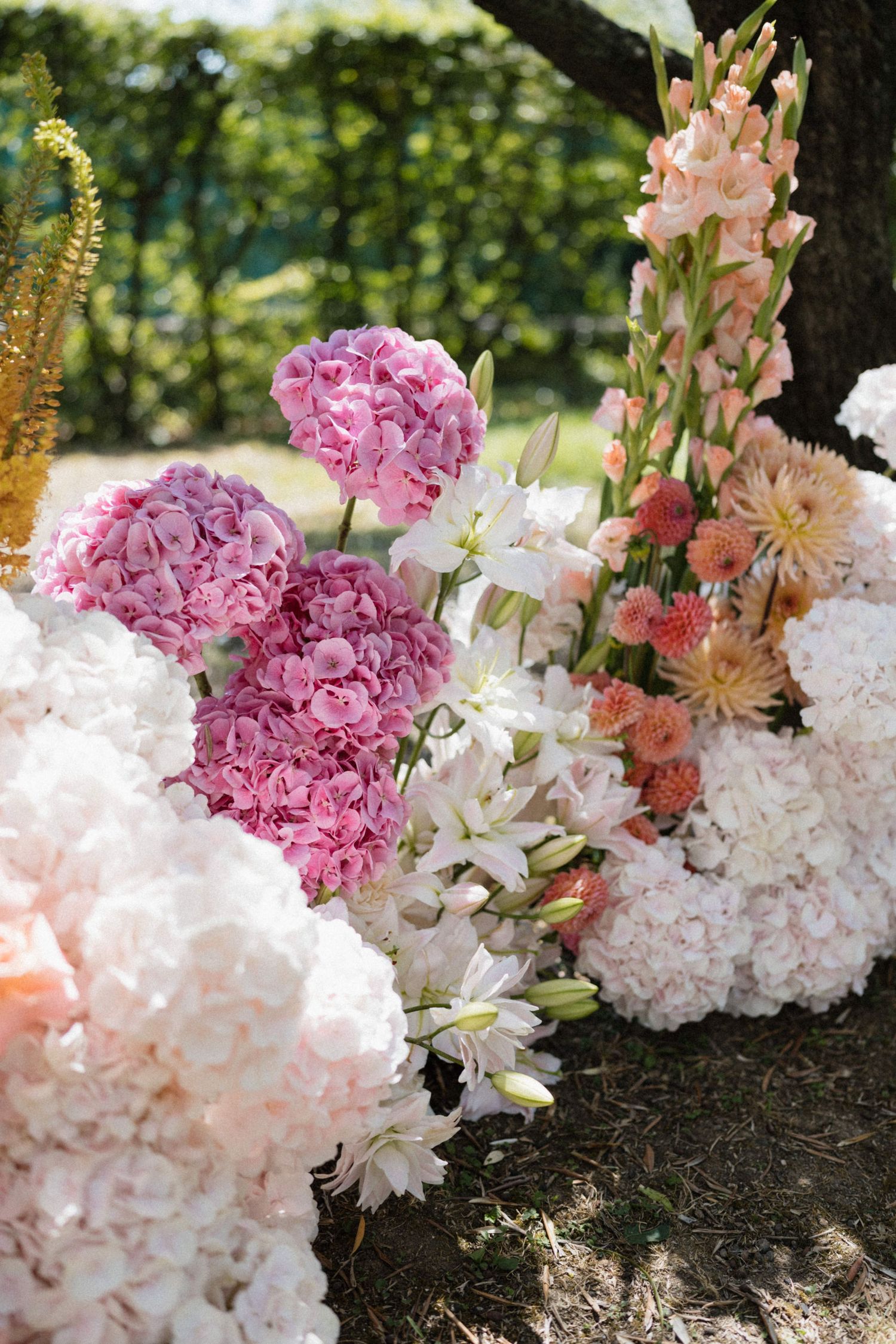 Lush arrangement of pink hydrangeas, white lilies, gladiolus, and peach dahlias displayed outdoors at a garden wedding ceremony.