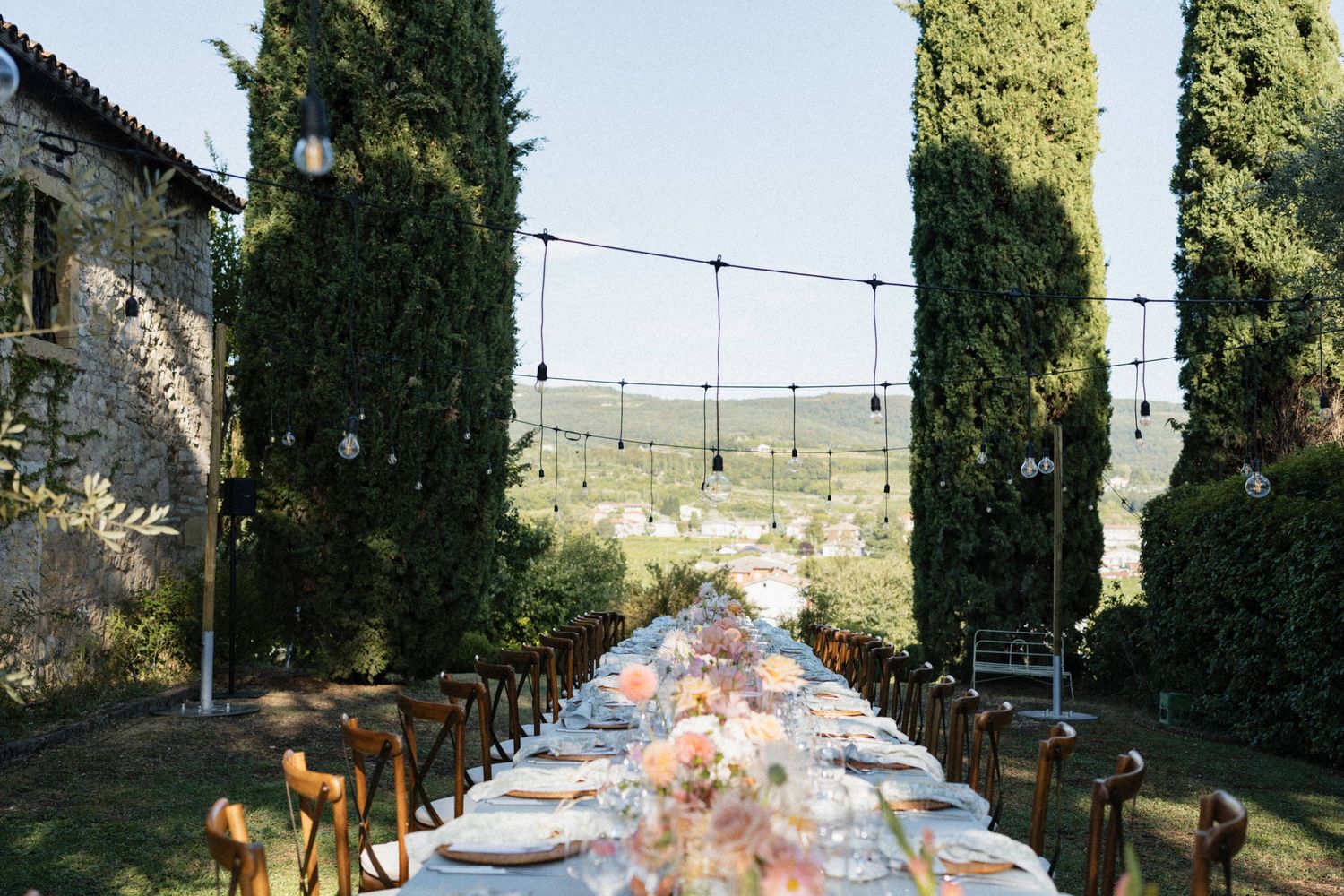 Elegant outdoor wedding reception with long table, floral centerpieces, cypress trees, and string lights in Tuscany.