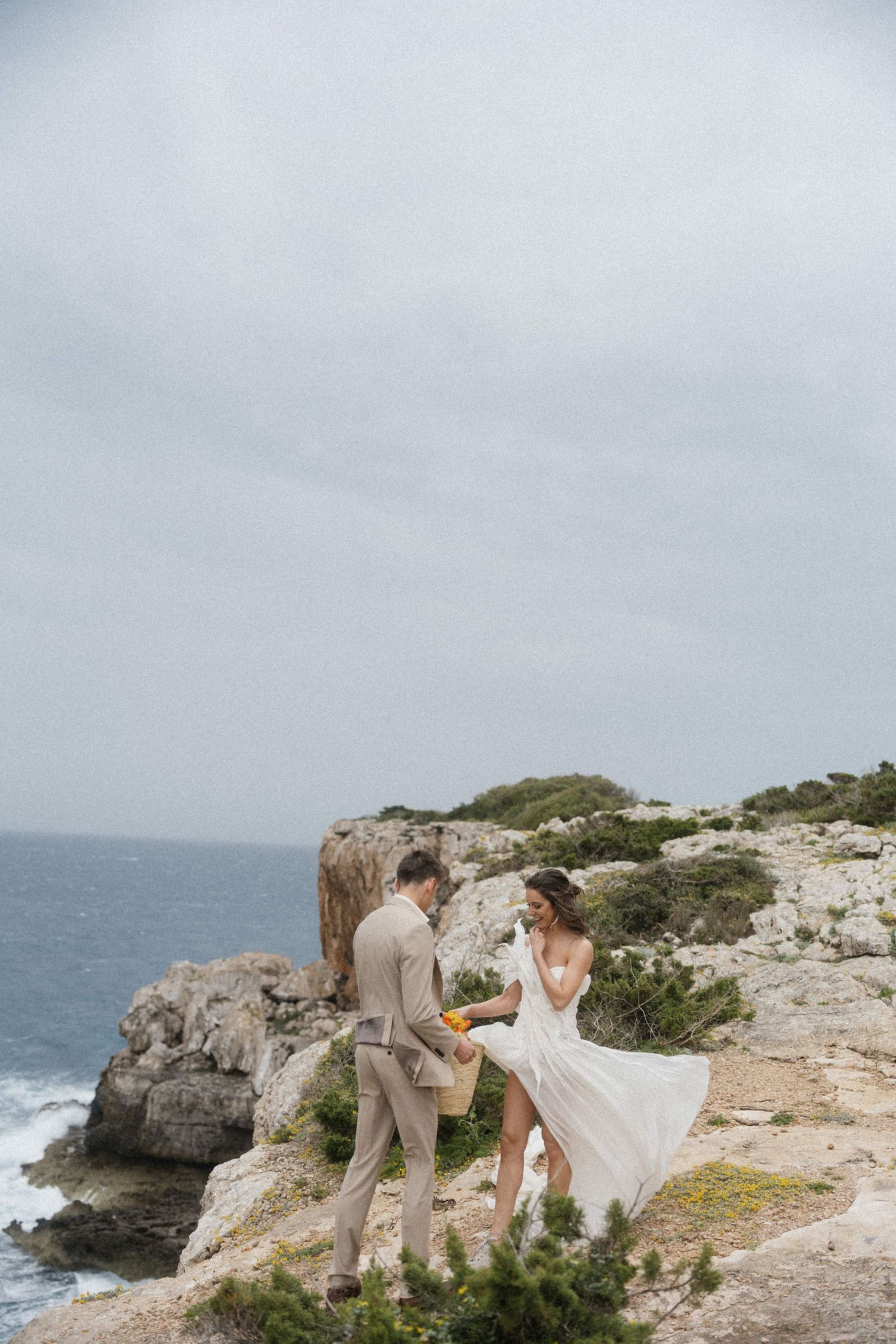 Bride and groom standing on rocky seaside cliffs, wedding dress flowing in the wind.