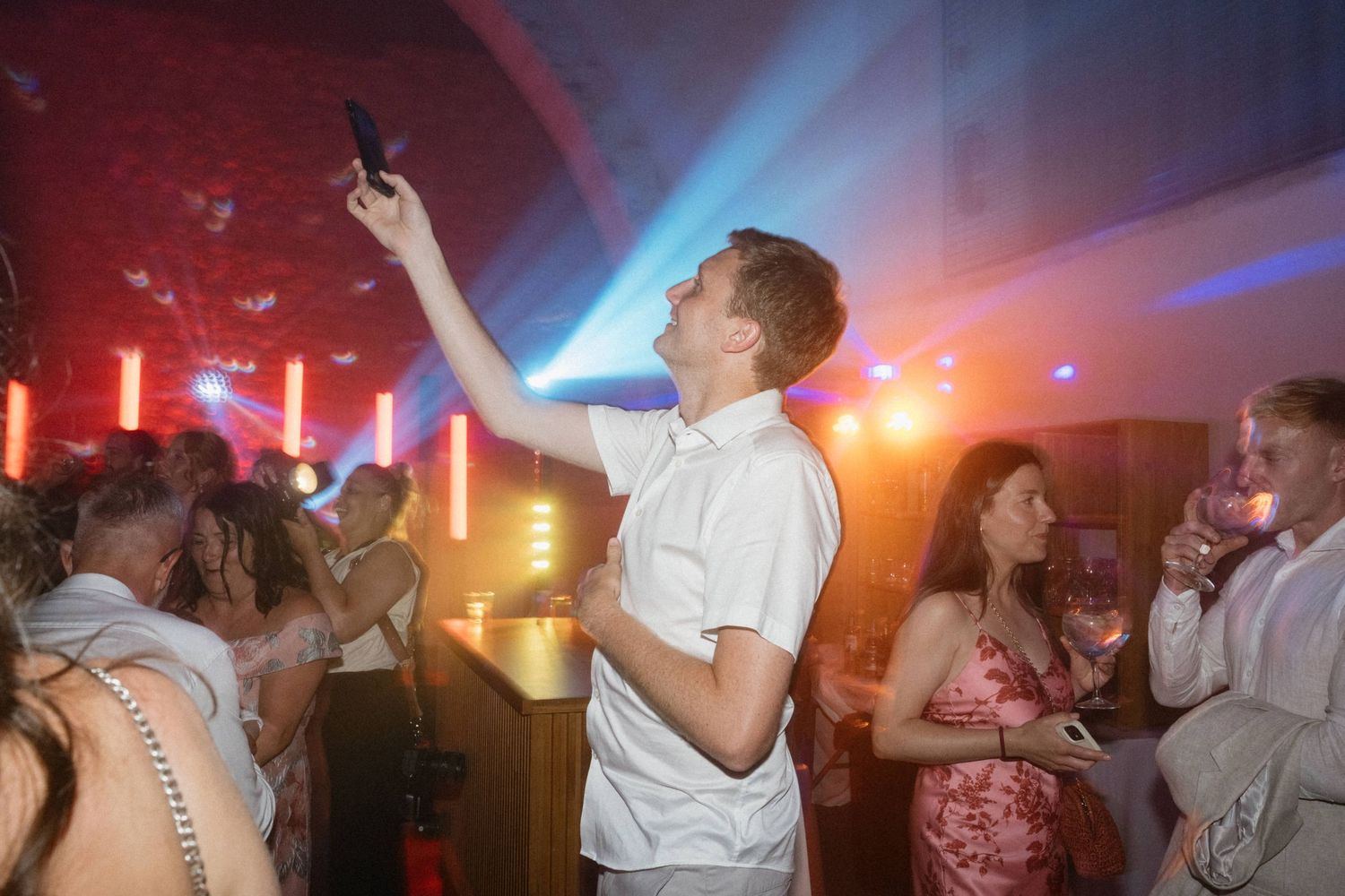 Man taking selfie with phone at crowded nightclub party with colorful neon lights and dancing guests.