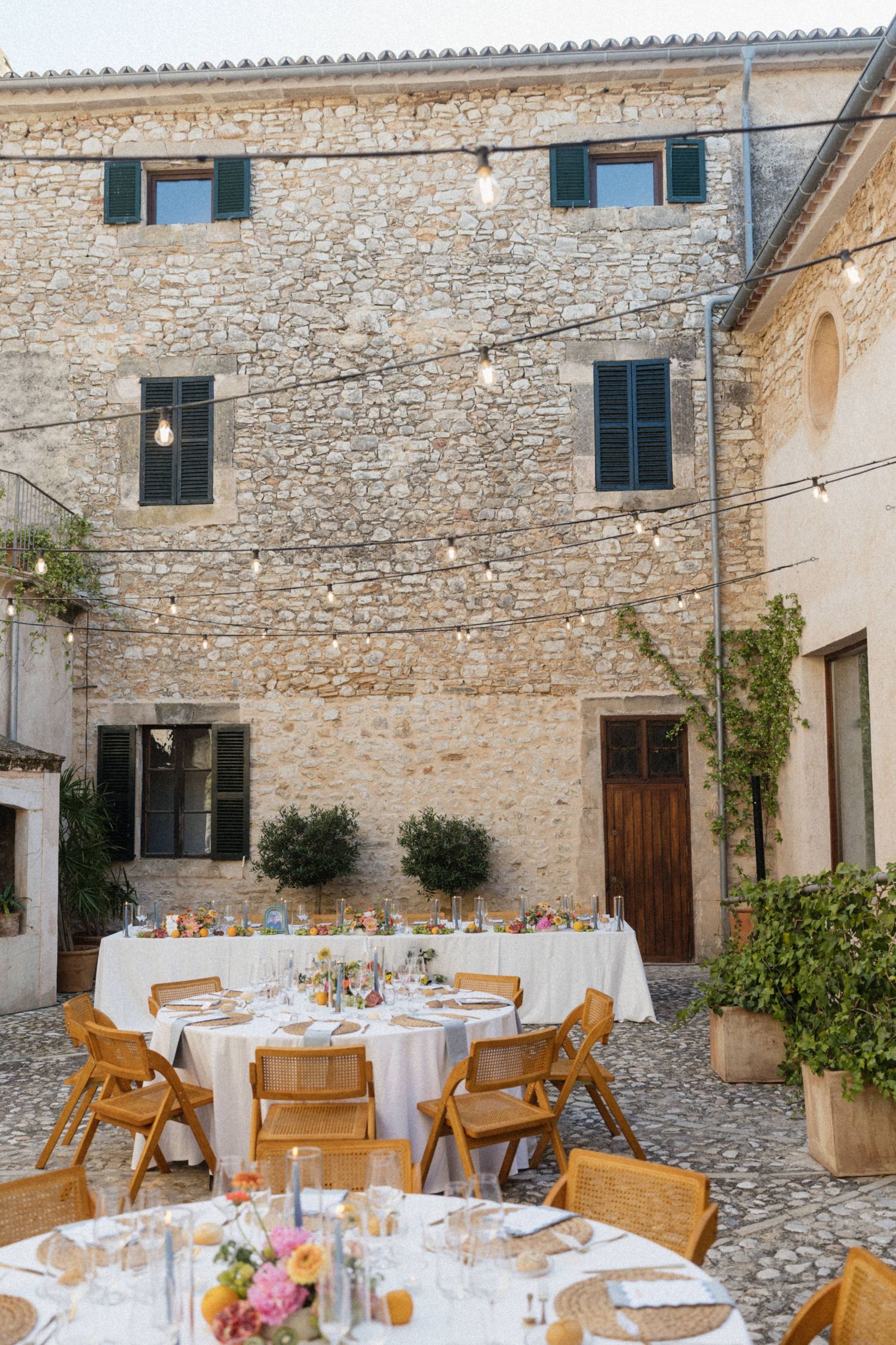 Outdoor wedding reception in a rustic stone courtyard with round tables, wooden chairs, and string lights.