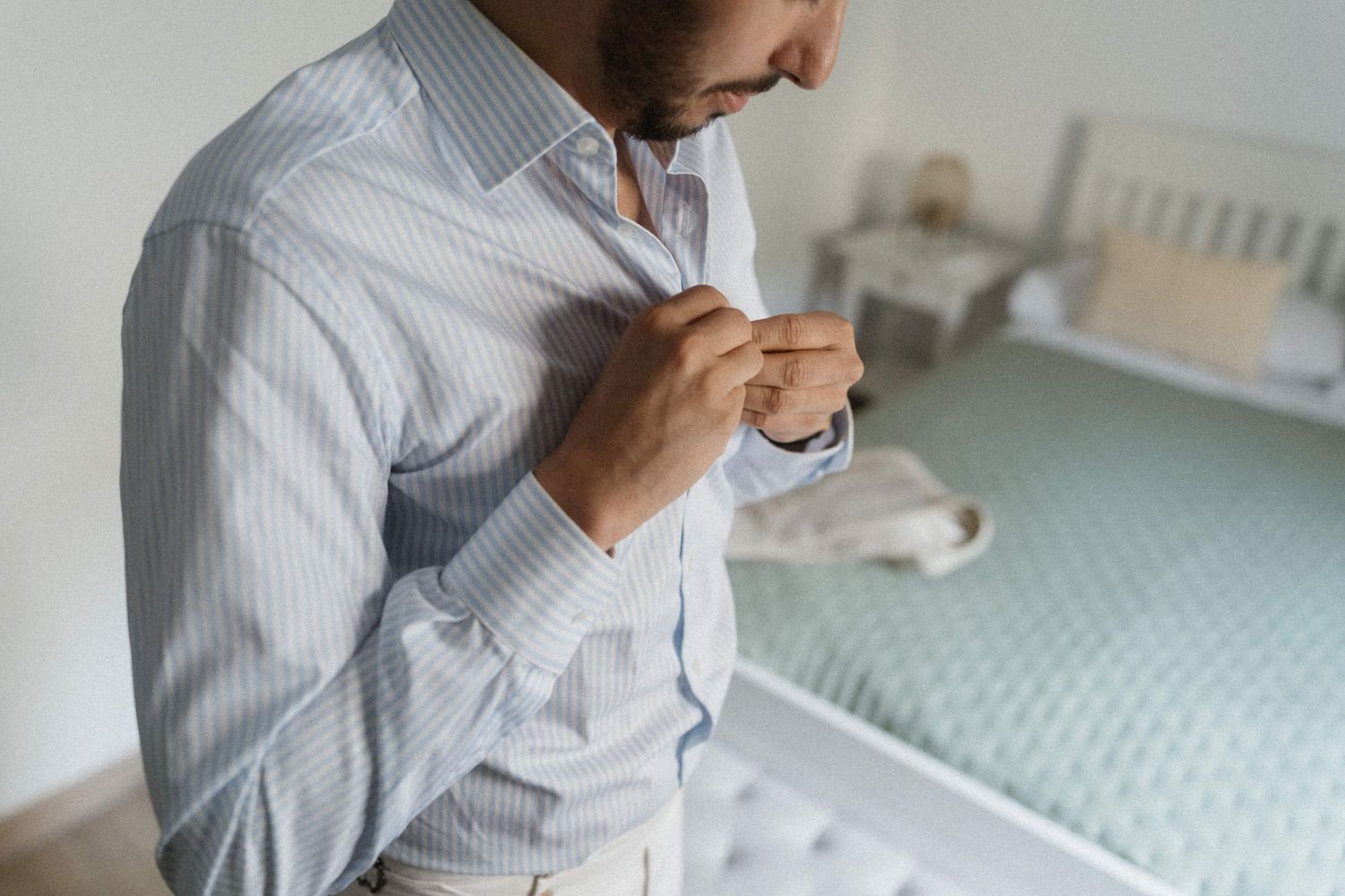 A man in a light blue striped shirt buttoning his collar in a bright bedroom while getting ready for a wedding.