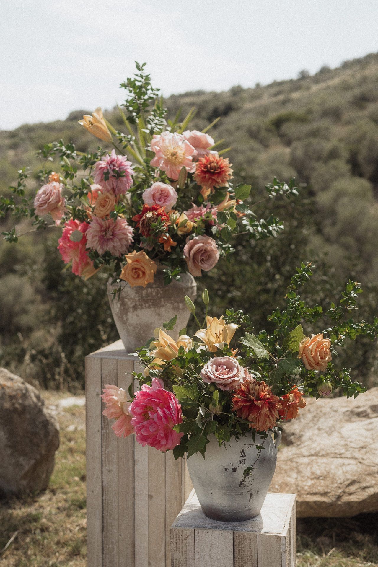 Two rustic vases with lush pink, peach, and coral floral arrangements displayed on white pedestals against a natural hillside backdrop.