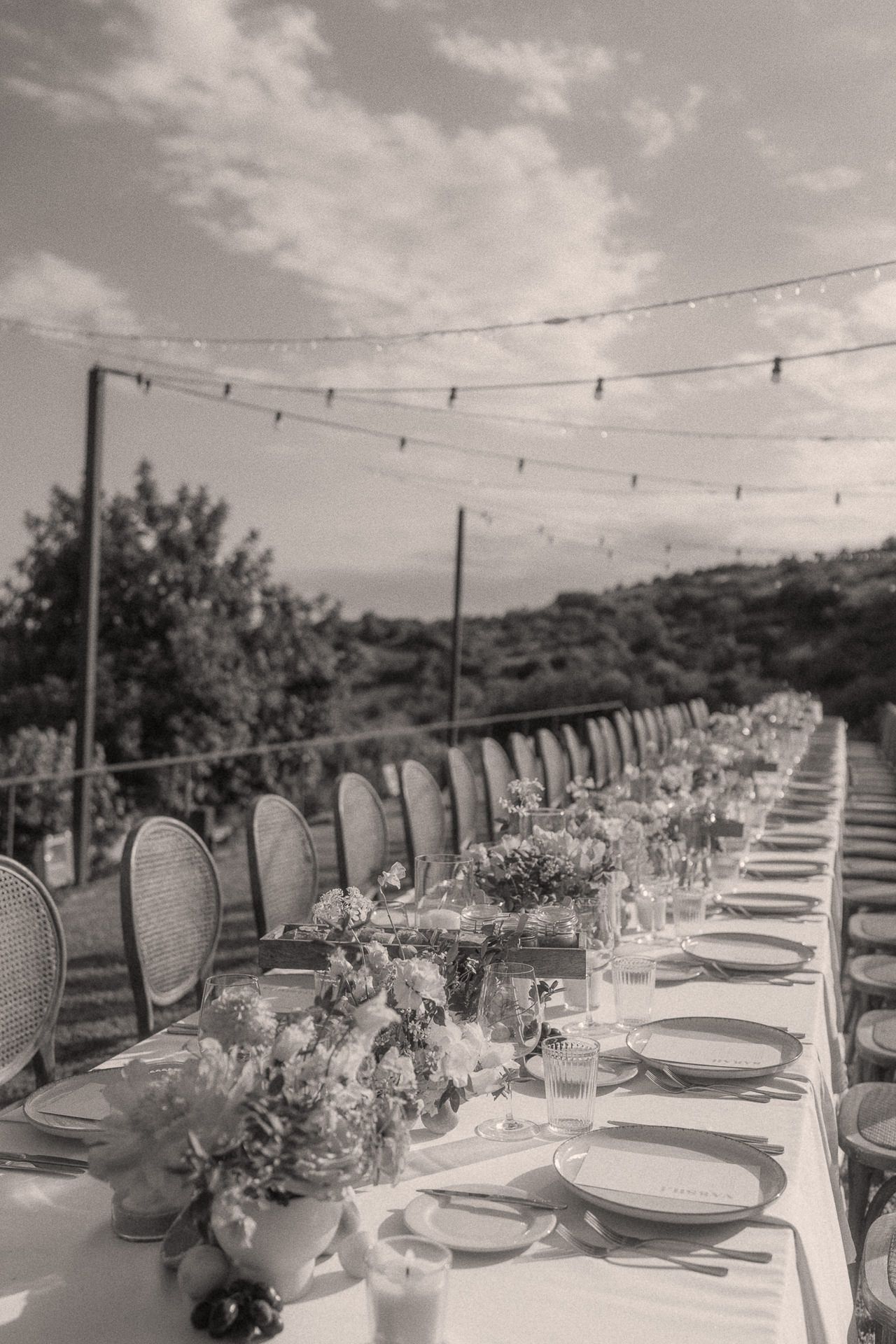 Black and white outdoor wedding reception table set with floral centerpieces, string lights, and elegant place settings.