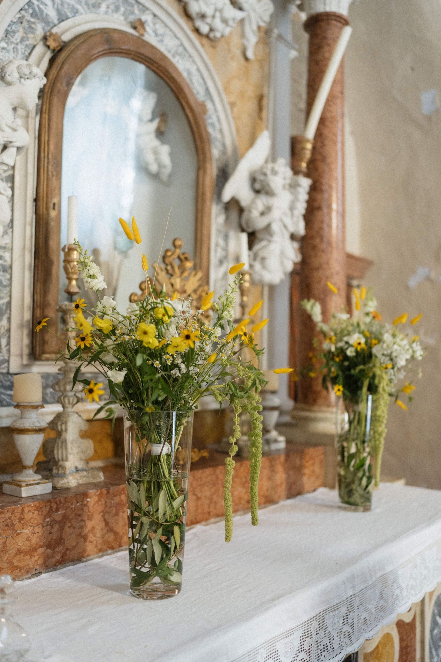 Glass vases with yellow wildflowers adorn a white linen-covered altar in an ornate church with marble columns.