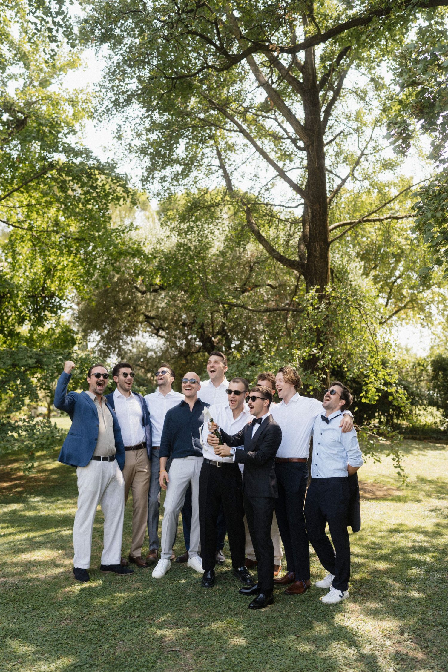 Groom in black tuxedo celebrating with groomsmen outdoors under green trees on sunny wedding day.