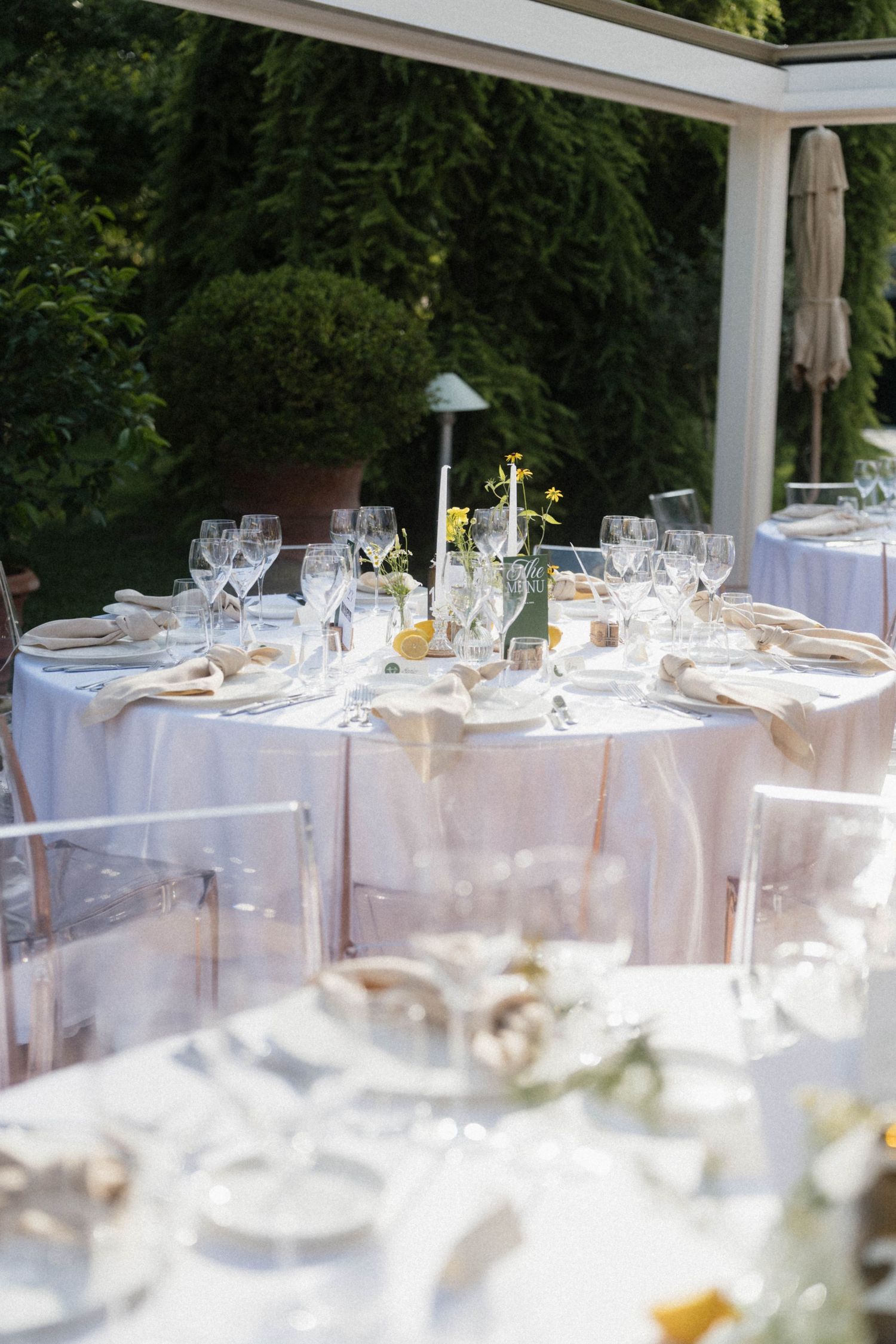 Elegant outdoor wedding reception tables with white linens, crystal glasses, and floral centerpieces under a pergola.