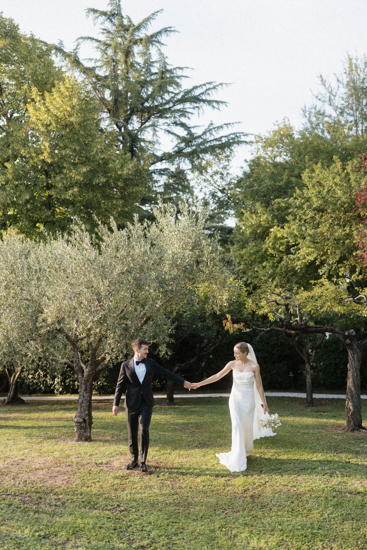 Bride and groom hold hands walking through sunlit garden with olive and cedar trees at Italian villa wedding.