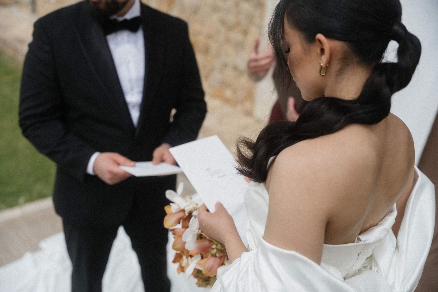 Groom in black tuxedo and bride in white dress exchanging written vows at outdoor ceremony.