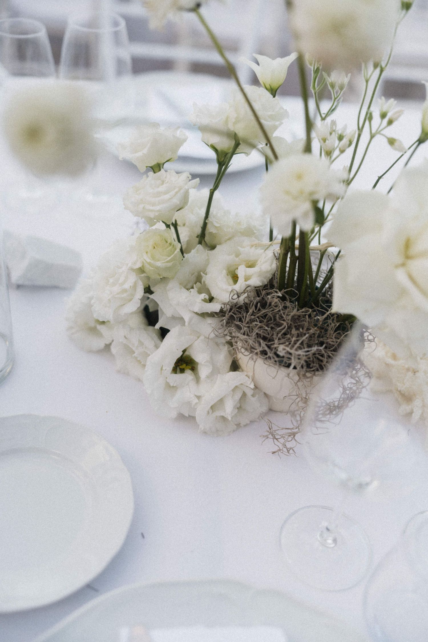 White wedding table centerpiece featuring roses, lisianthus, and wildflowers arranged with moss in a natural setting.