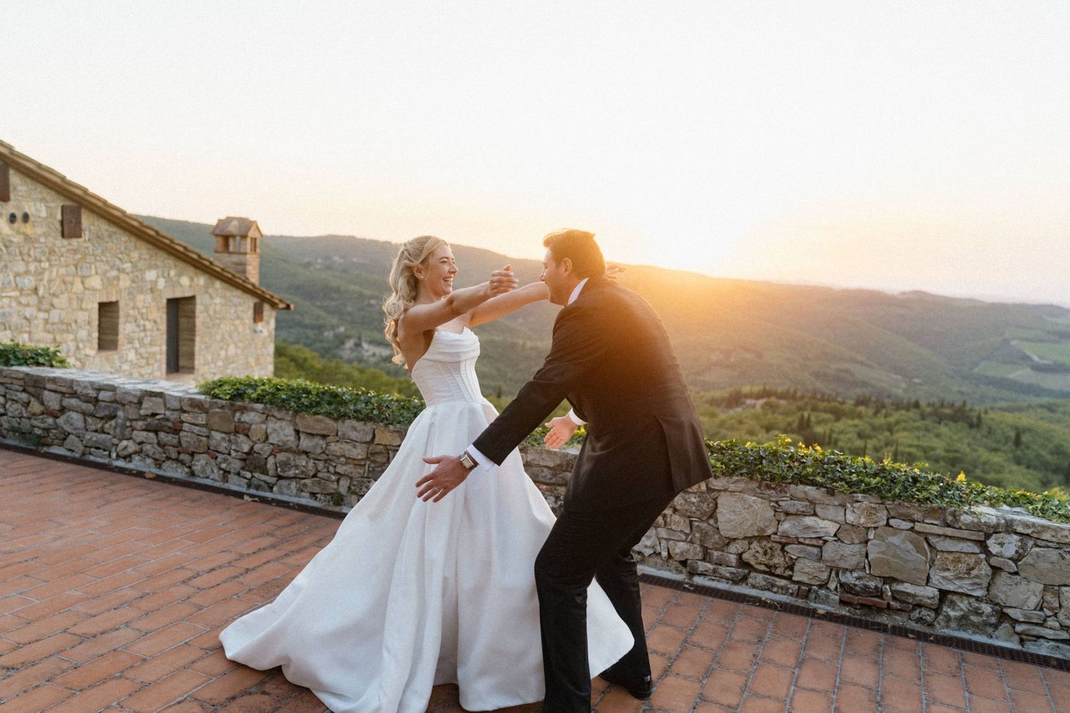 Bride and groom dancing joyfully on a terrace with a stunning Tuscan sunset backdrop.