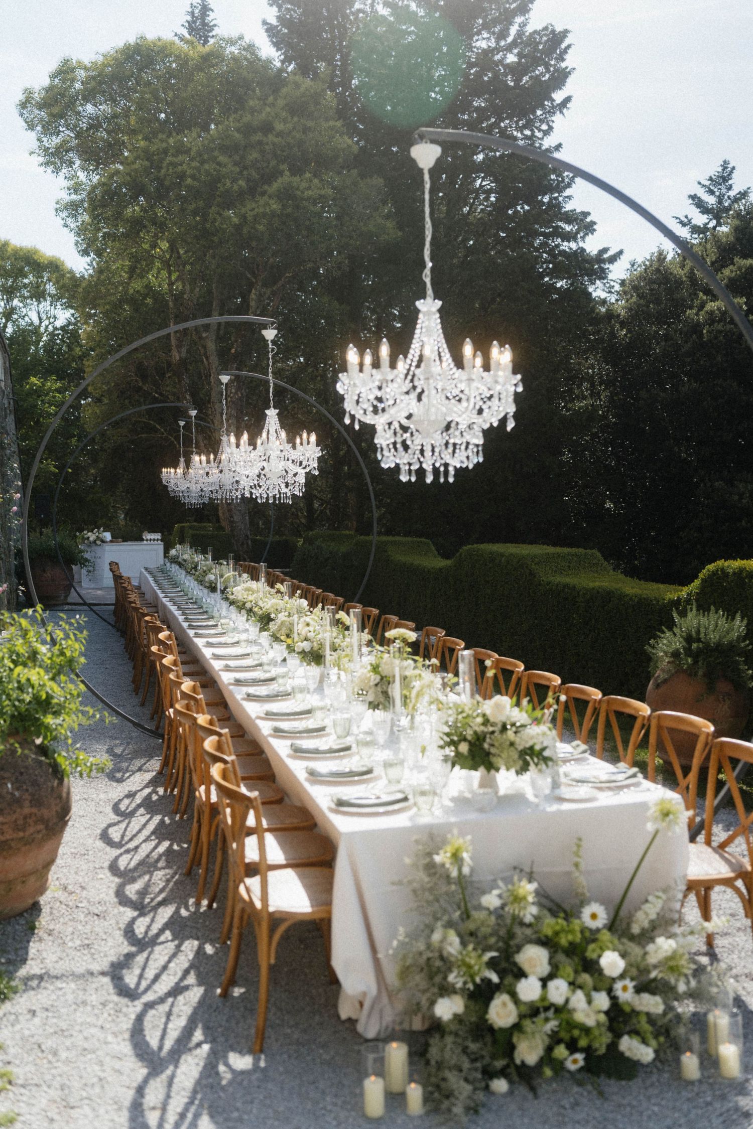 Elegant outdoor wedding reception with long table, crystal chandeliers, wood chairs, and white floral centerpieces.