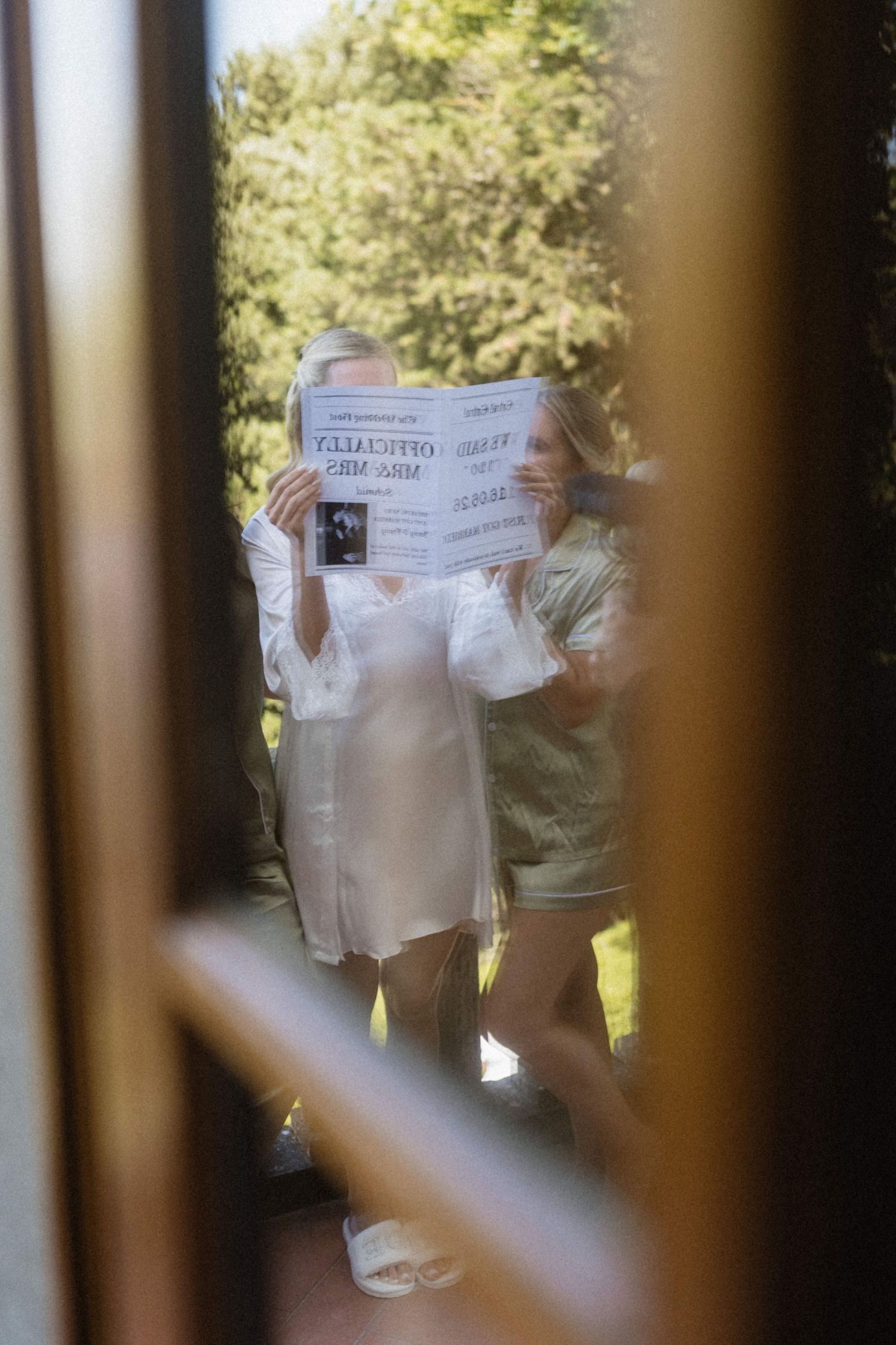 Bride in white dress reads a newspaper outdoors, reflected in a window at a wedding venue.