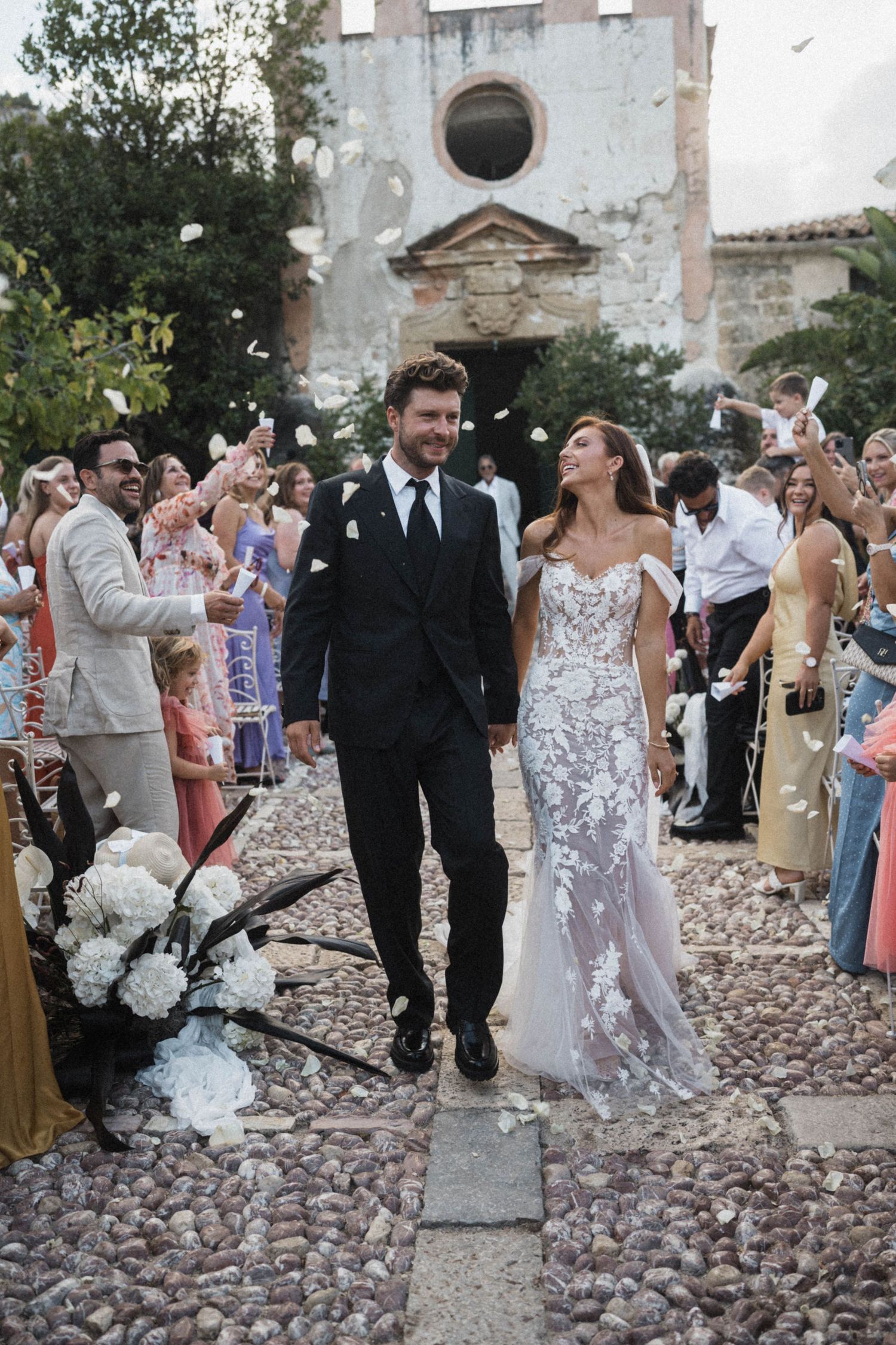 Newlyweds walk down aisle as guests toss white flower petals in celebration.