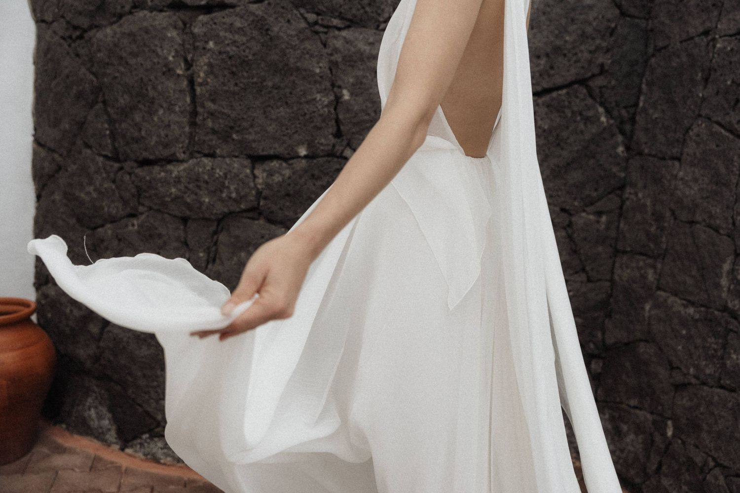 Woman in flowing white bridal gown holding dress hem against dark volcanic stone wall.