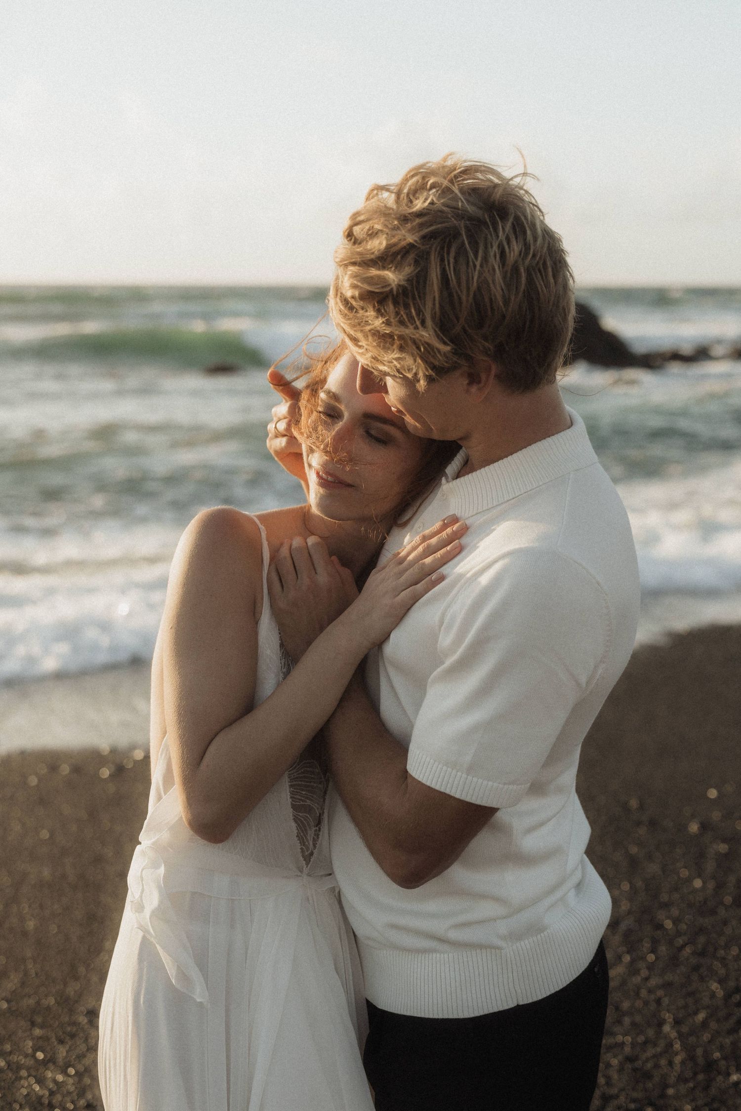 Couple embracing tenderly on black sand beach at sunset with ocean waves behind them.