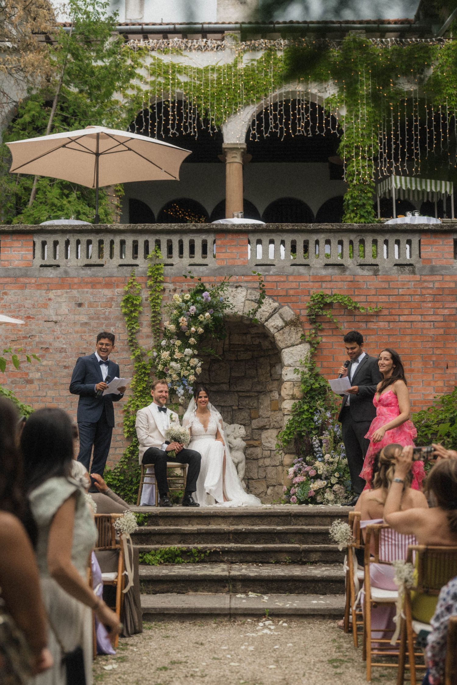 Bride and groom seated at an outdoor wedding ceremony with a floral arch against a rustic brick wall.