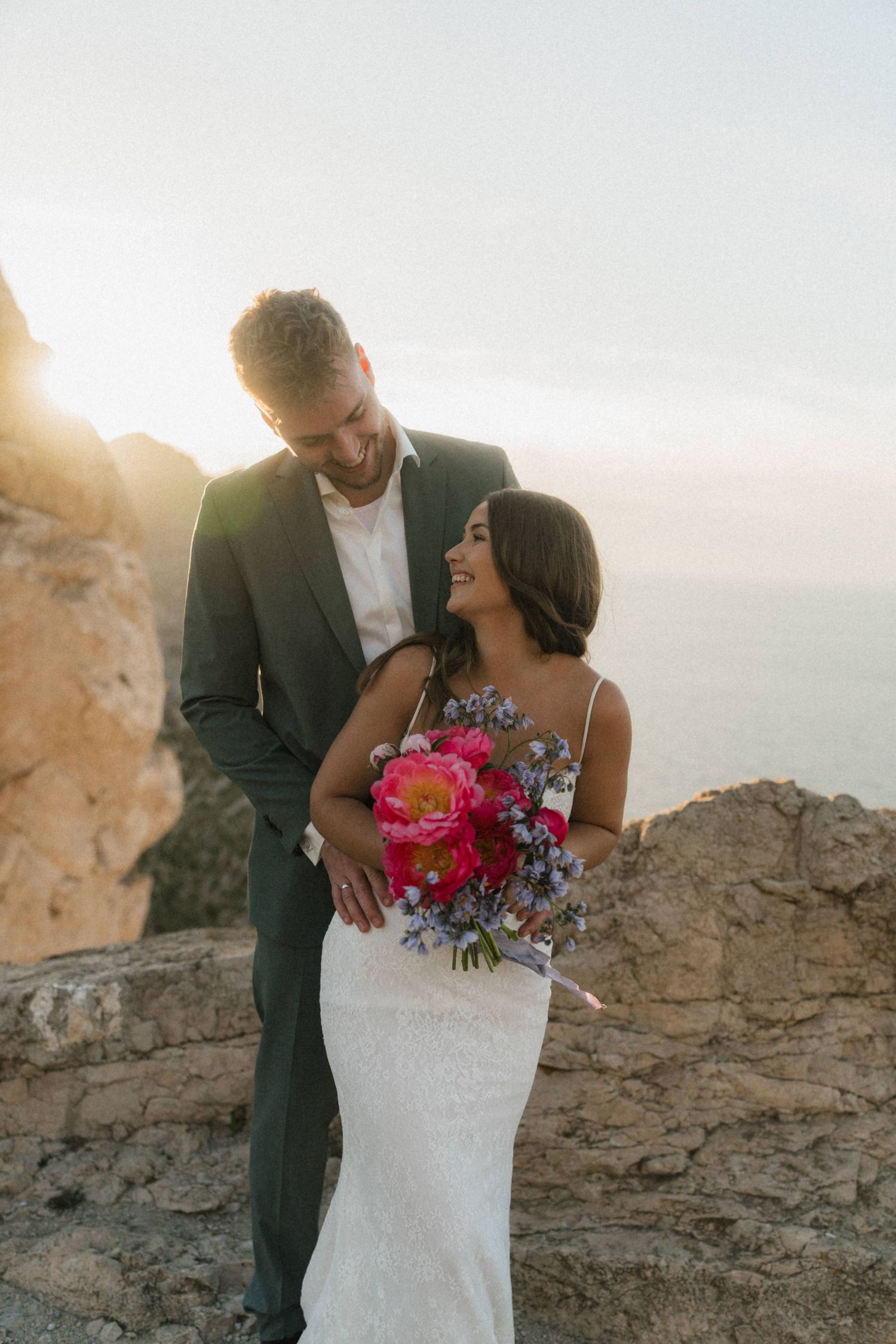 Bride and groom embracing on rocky cliffside at sunset, bride holding vibrant pink and blue bouquet with ocean backdrop.