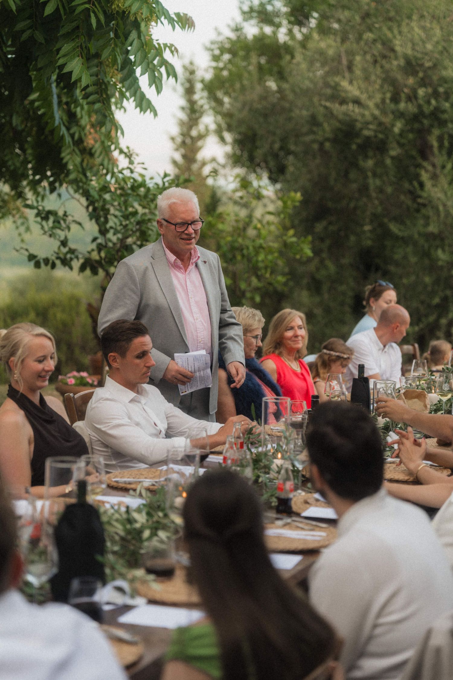 Elderly man in grey blazer gives speech at elegant outdoor wedding reception dinner table.
