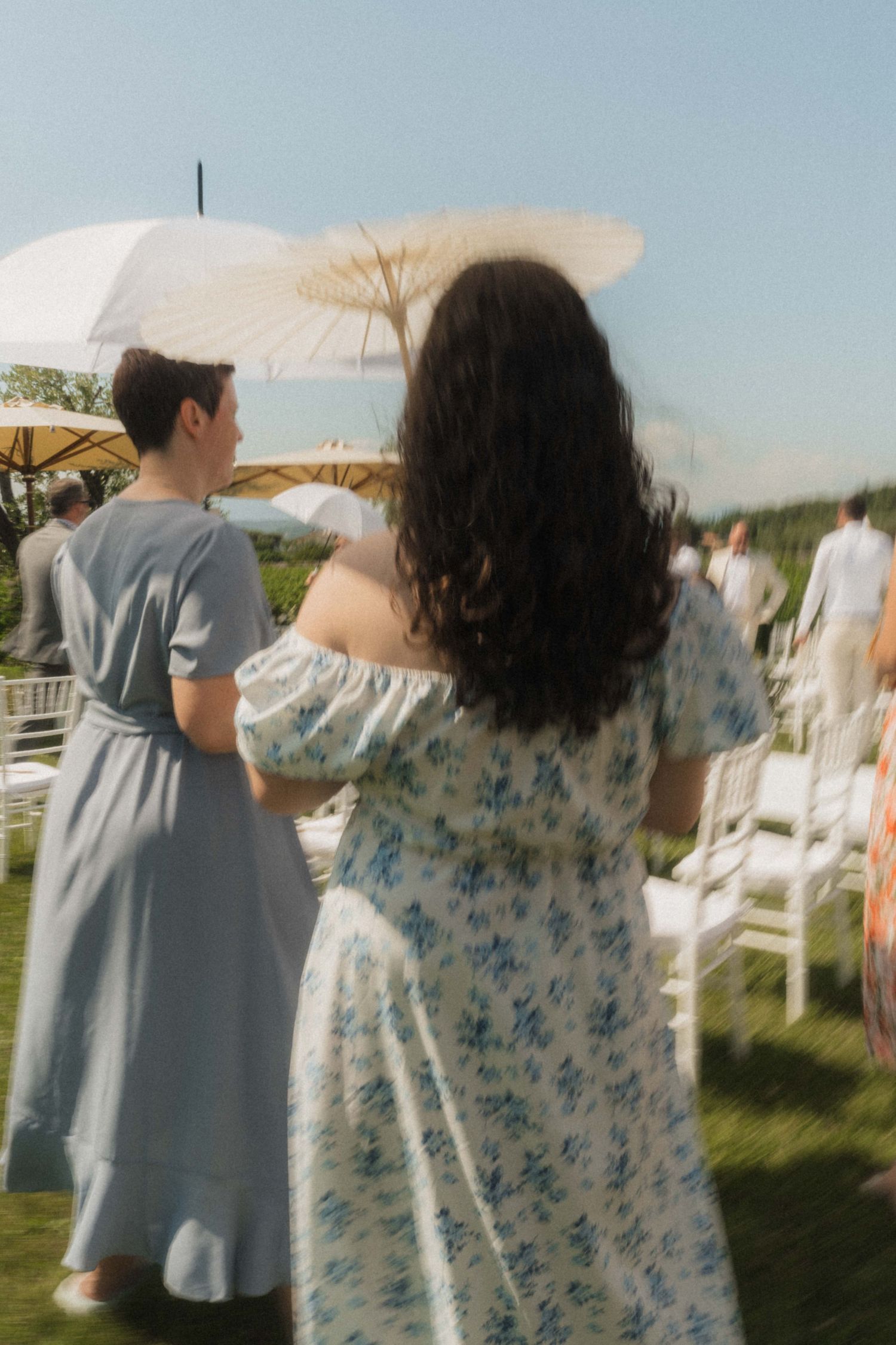 Wedding guests in floral and blue dresses holding parasols at an outdoor summer ceremony.