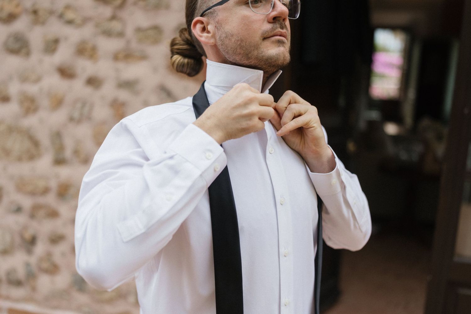 Groom in a white dress shirt and glasses adjusts his black bow tie while getting ready for his wedding beside a rustic stone wall.