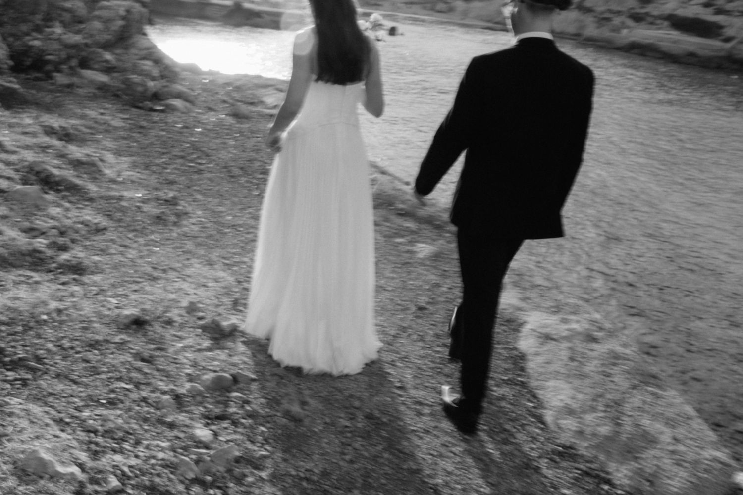 Black and white photo of a bride in a flowing gown and a groom in a suit walking away along a rocky coastal path.