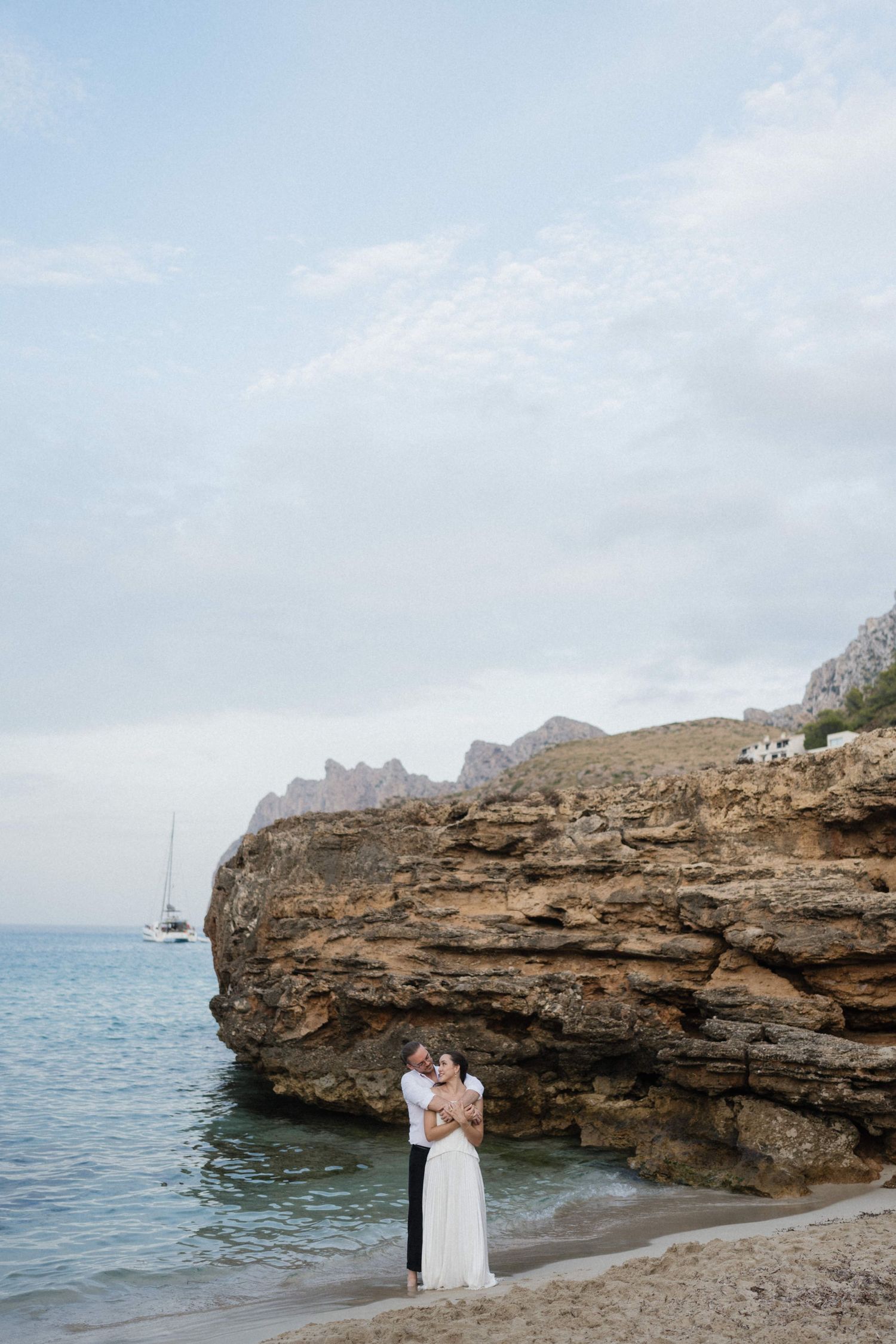 Couple hugging on a secluded beach below rugged coastal cliffs, with a sailboat on the calm sea and cloudy sky behind them.