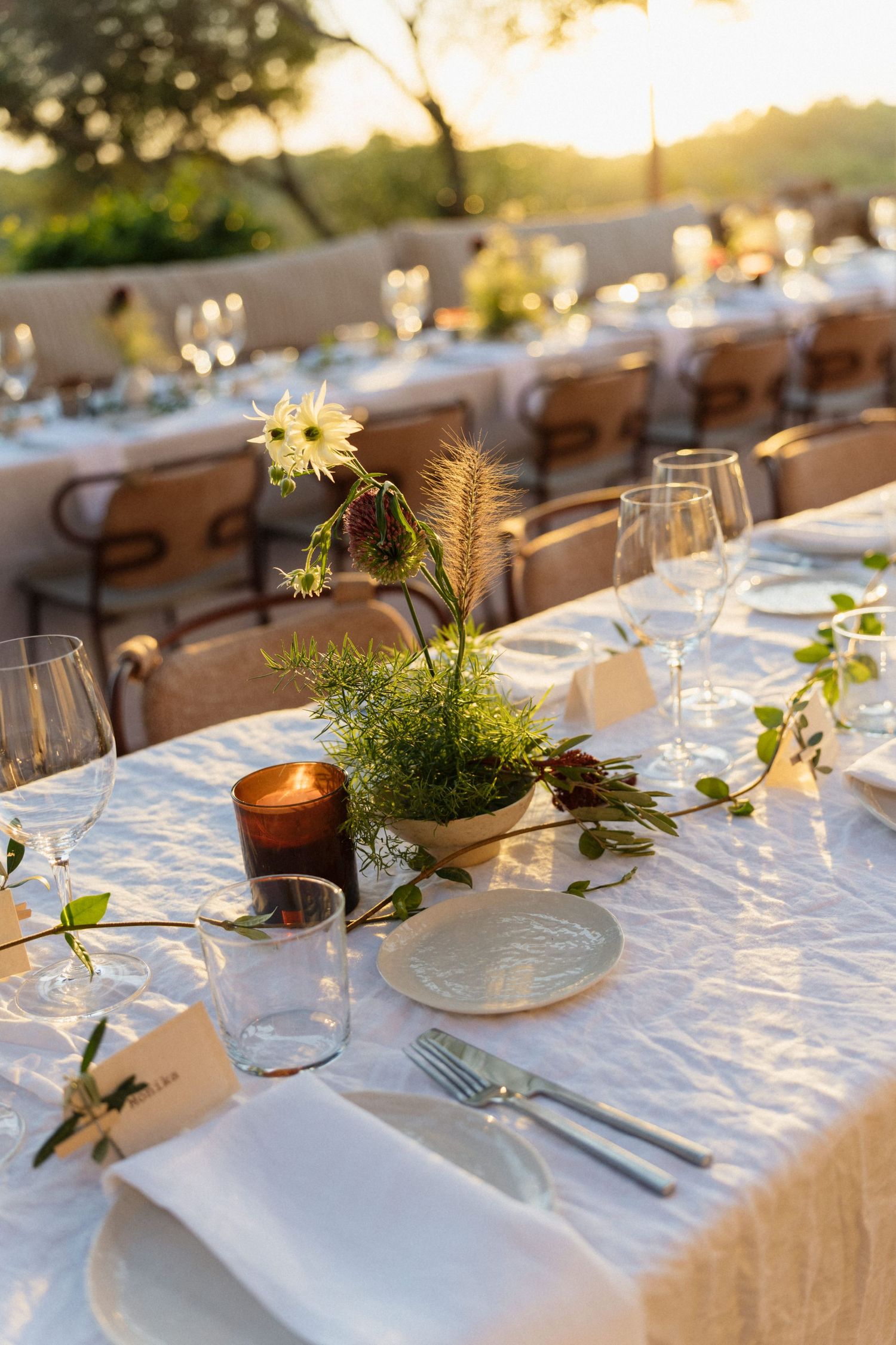 A long wedding reception table at sunset is set with white linens, wine glasses, candles, place cards and wildflower centerpieces.