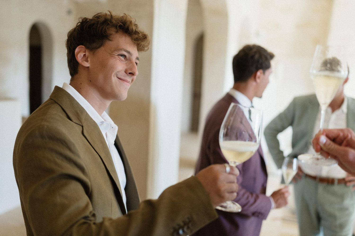 Smiling man in an olive blazer holds a glass of white wine at a stone-walled tasting event with guests behind him.