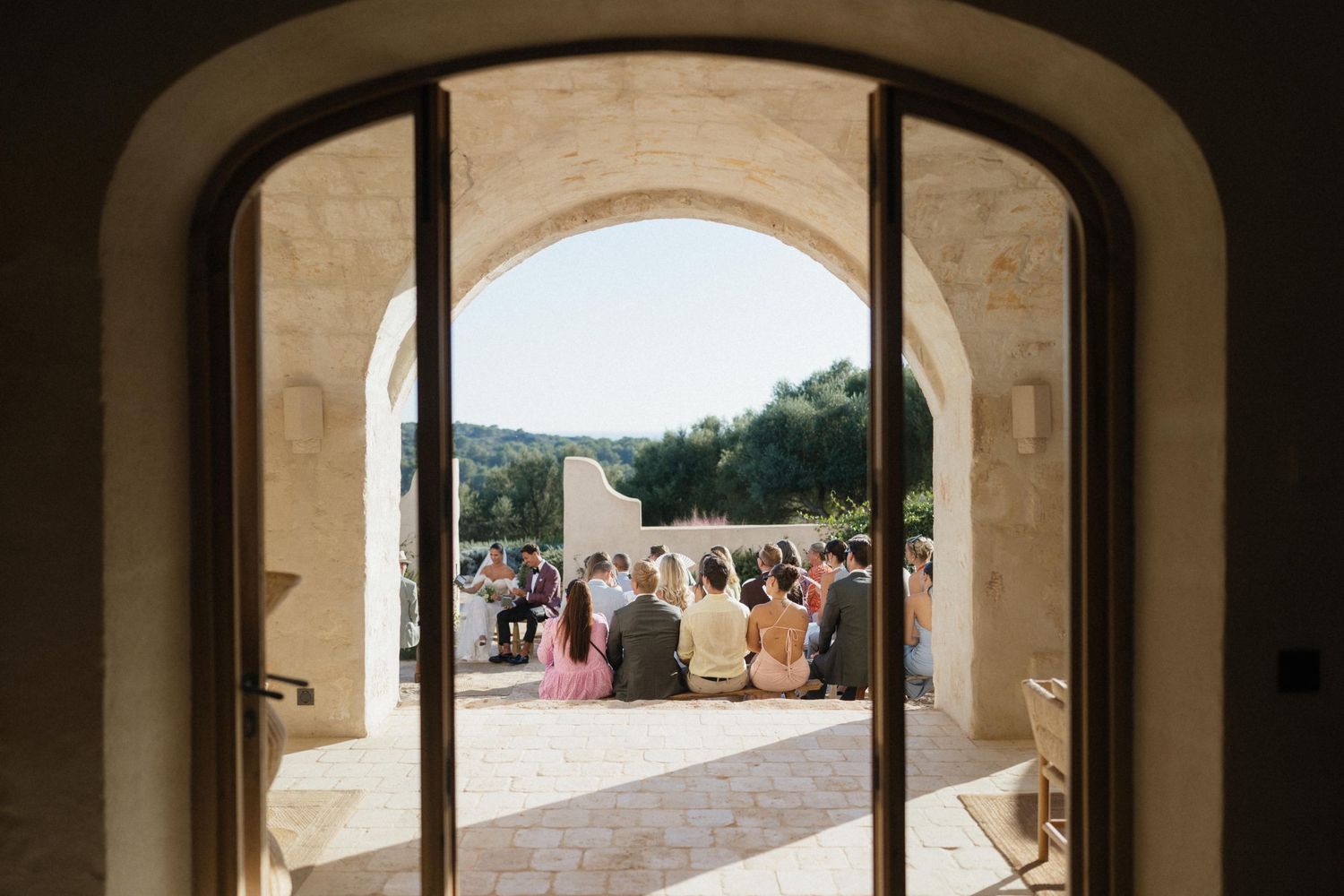 View through a stone archway of seated wedding guests gathered for an outdoor ceremony on a sunny terrace surrounded by trees.