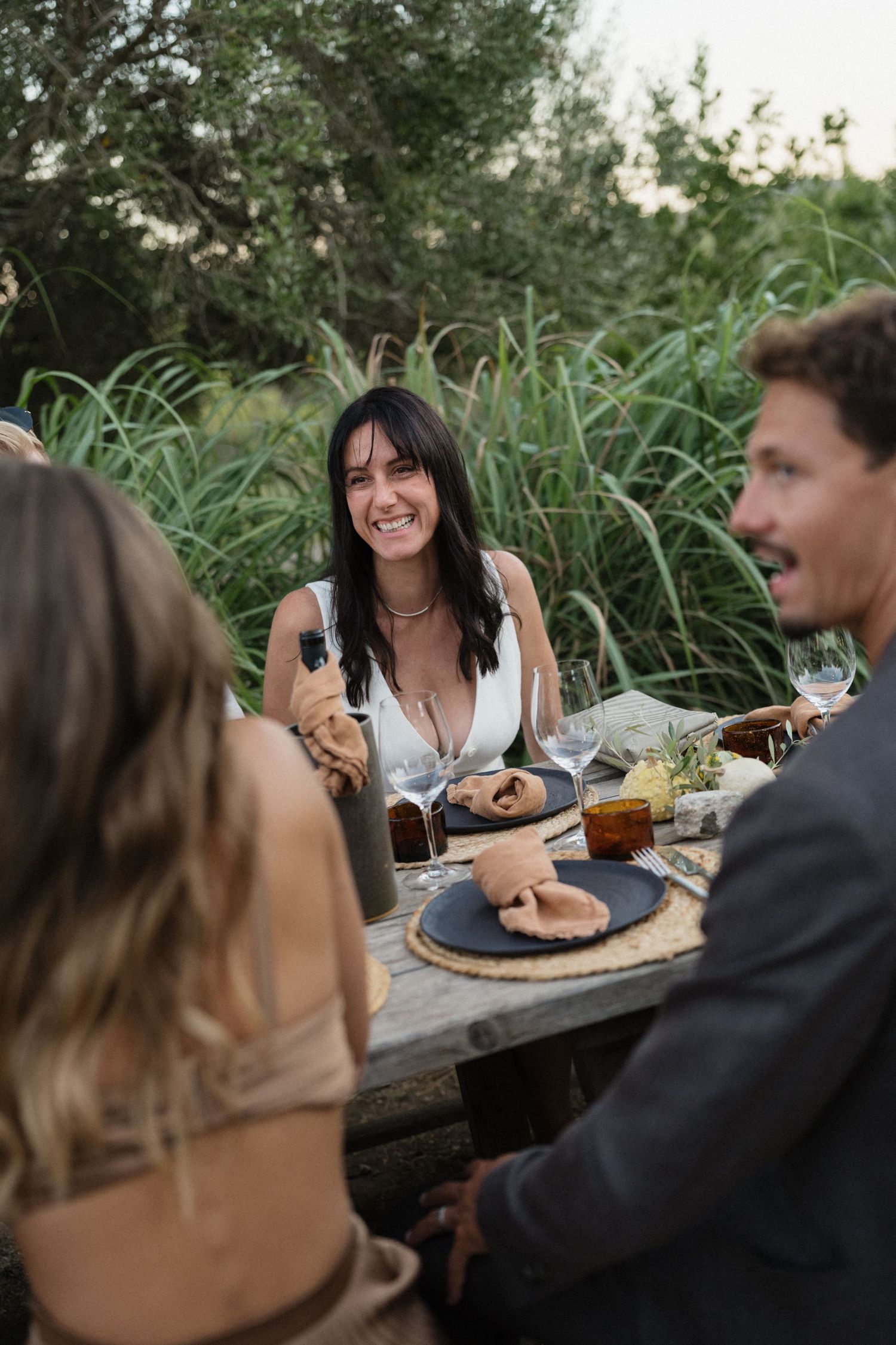 Group of friends laughing around a rustic outdoor table set with plates, napkins, and glasses among tall green grasses.