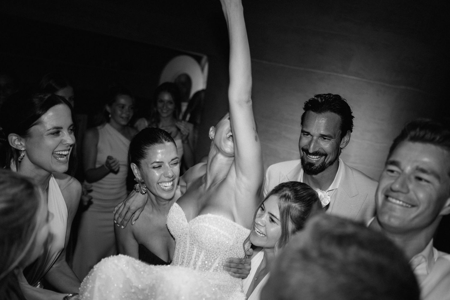 Black and white photo of joyful wedding guests lifting a bride in her gown on the dance floor.