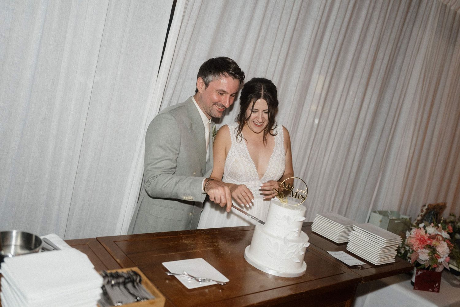 Bride and groom in grey suit cut white tiered wedding cake together at a wooden table indoors.
