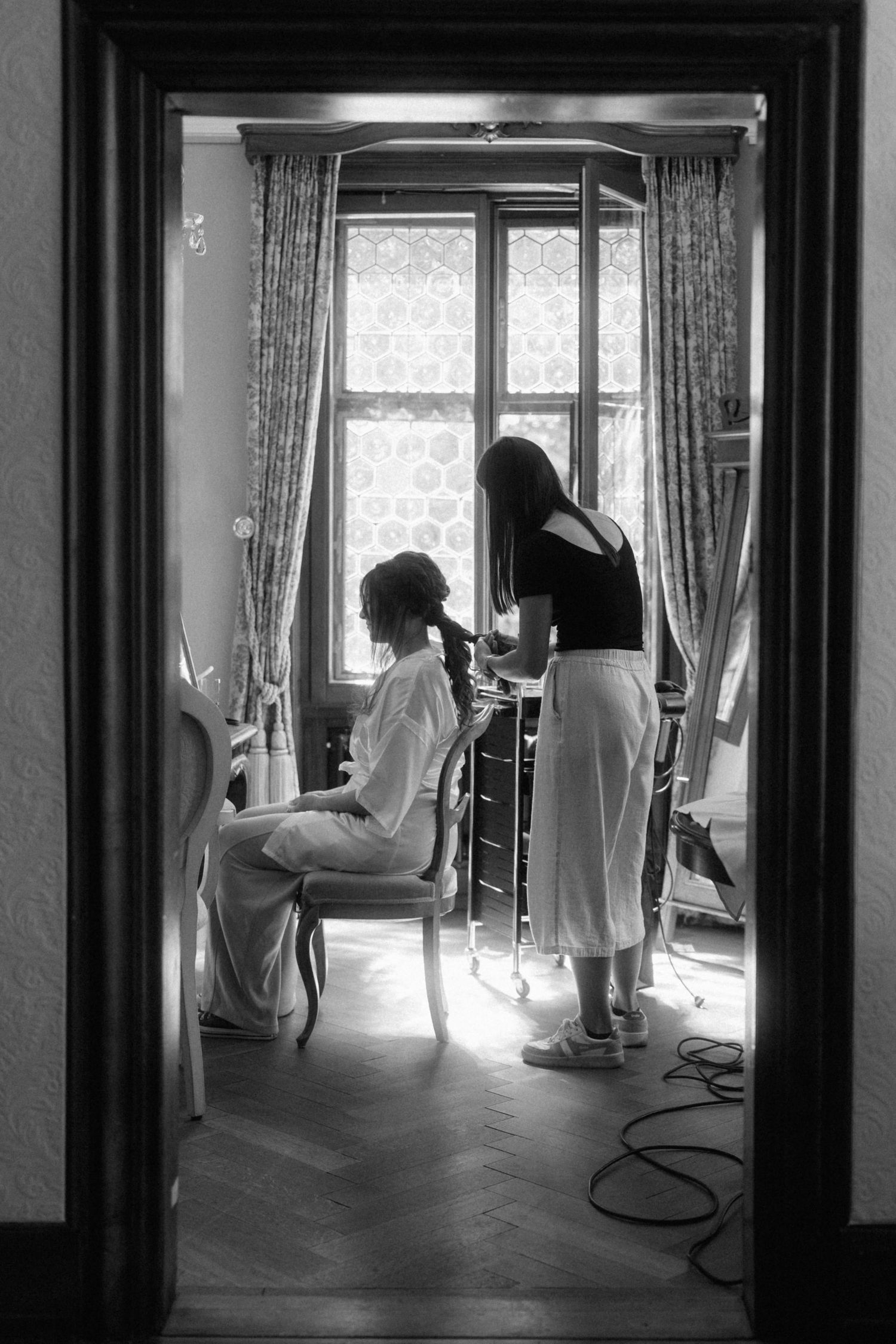 Black and white photo of a bride getting her hair done by a stylist in an elegant room with herringbone floors.