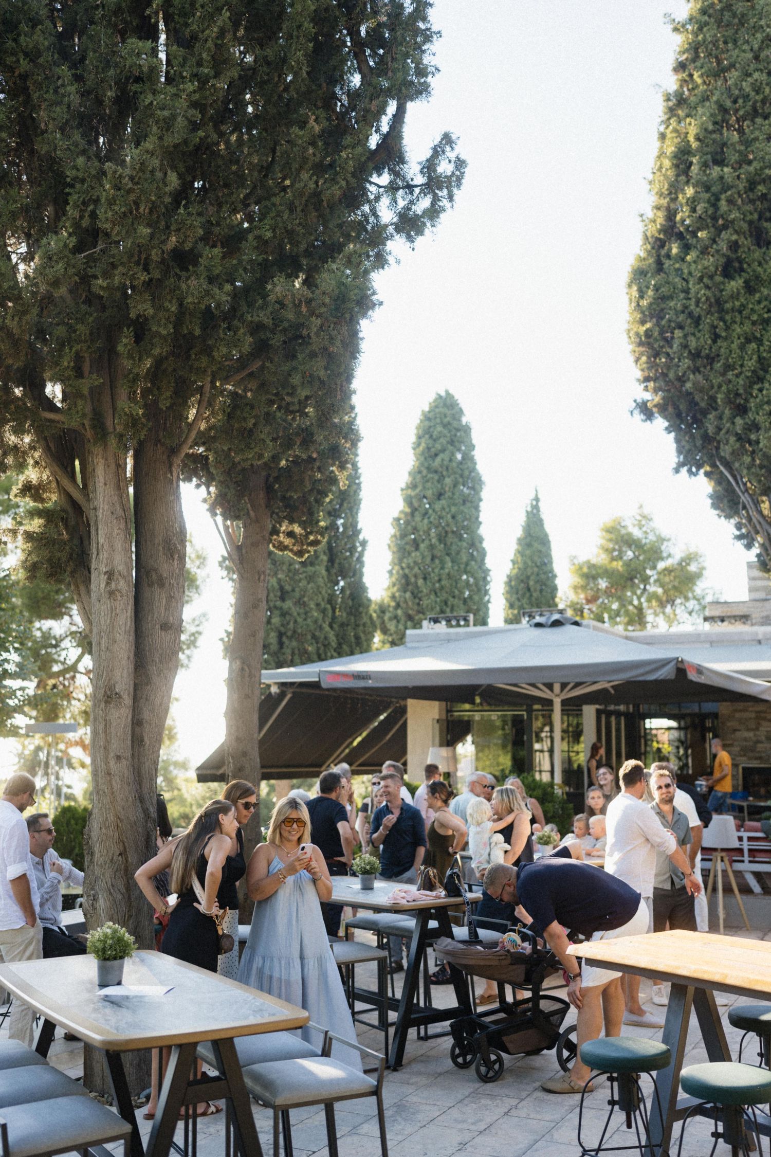 Guests mingle at an outdoor restaurant patio with tall cypress trees, umbrella shading, and modern tables on a sunny day.