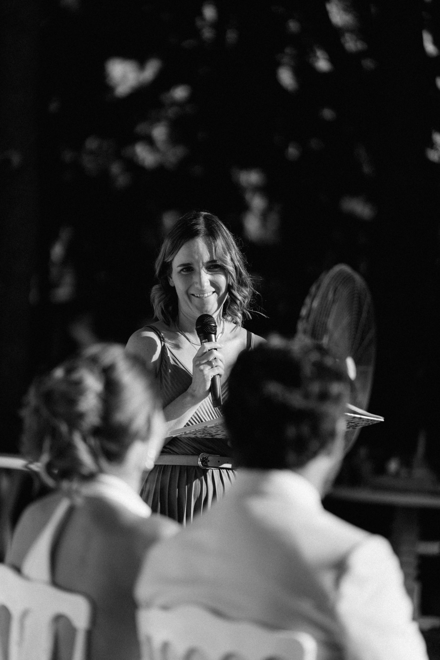 Black and white photo of a woman speaking into a microphone at an outdoor wedding ceremony with guests in the foreground.
