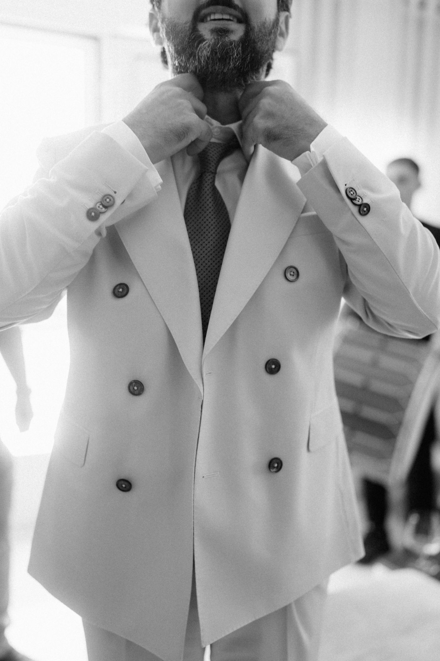 Groom in white double-breasted suit adjusting his tie, black and white photo.