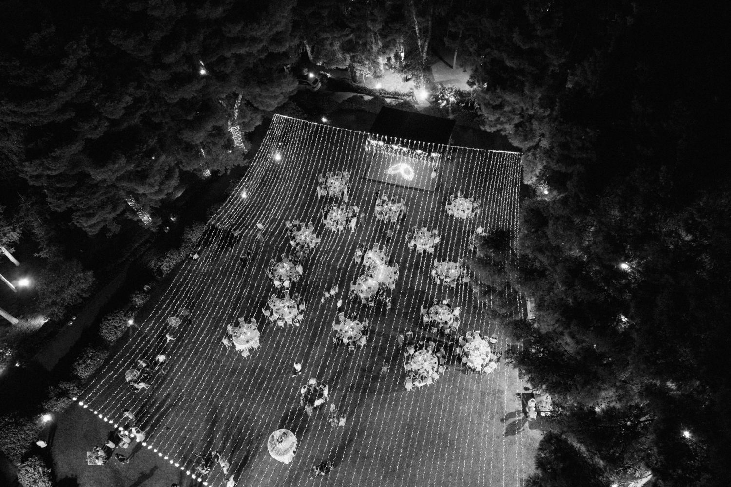 Black and white aerial night view of outdoor wedding reception with illuminated round tables and string lights.
