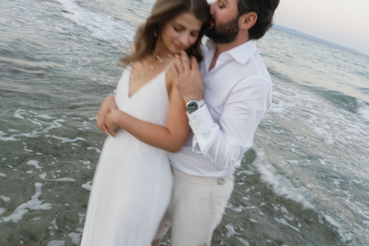 Romantic couple in white embracing on a beach shore at dusk with ocean waves.