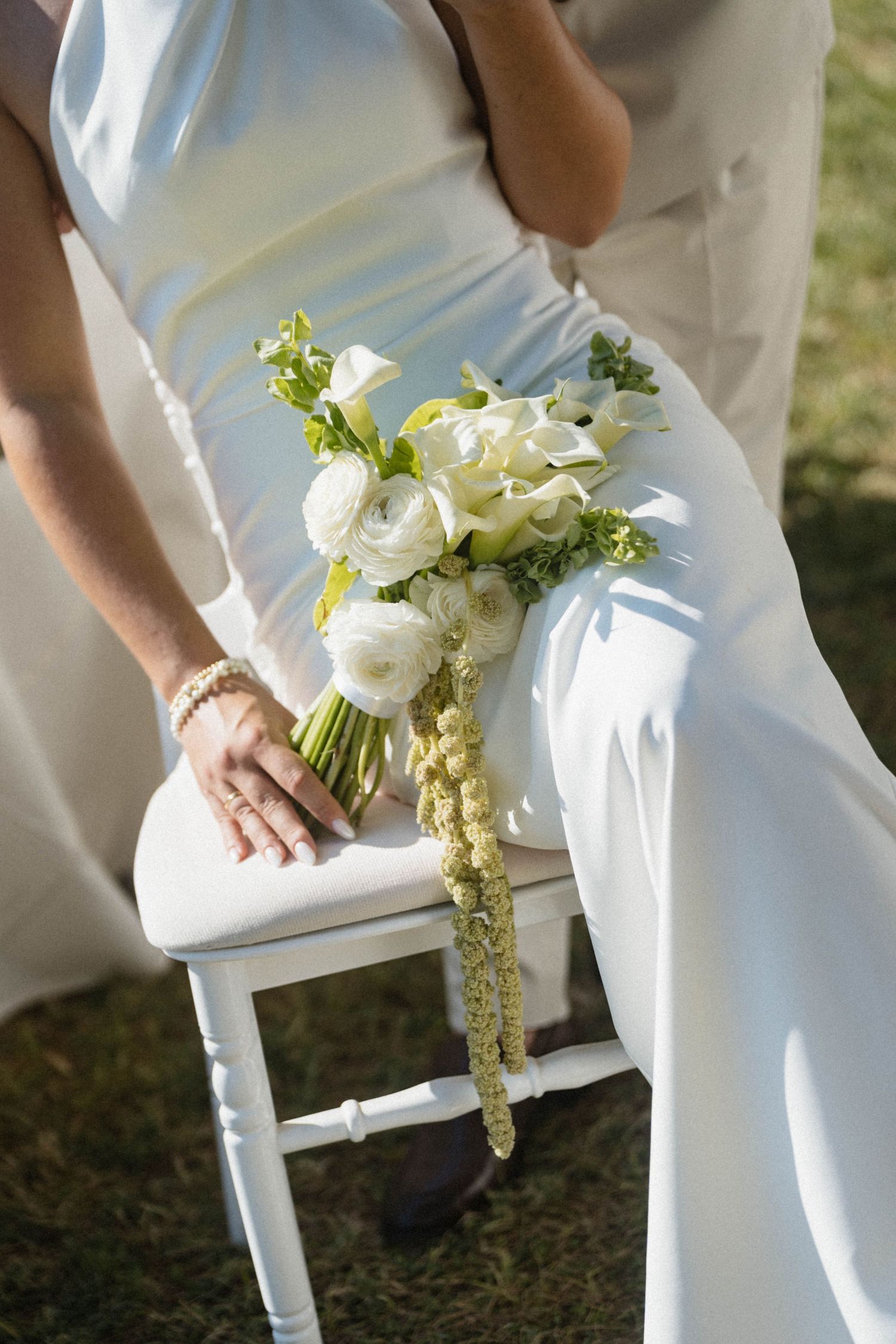 Bride in white gown sitting on chair holding elegant white wedding bouquet with ranunculus and calla lilies outdoors.