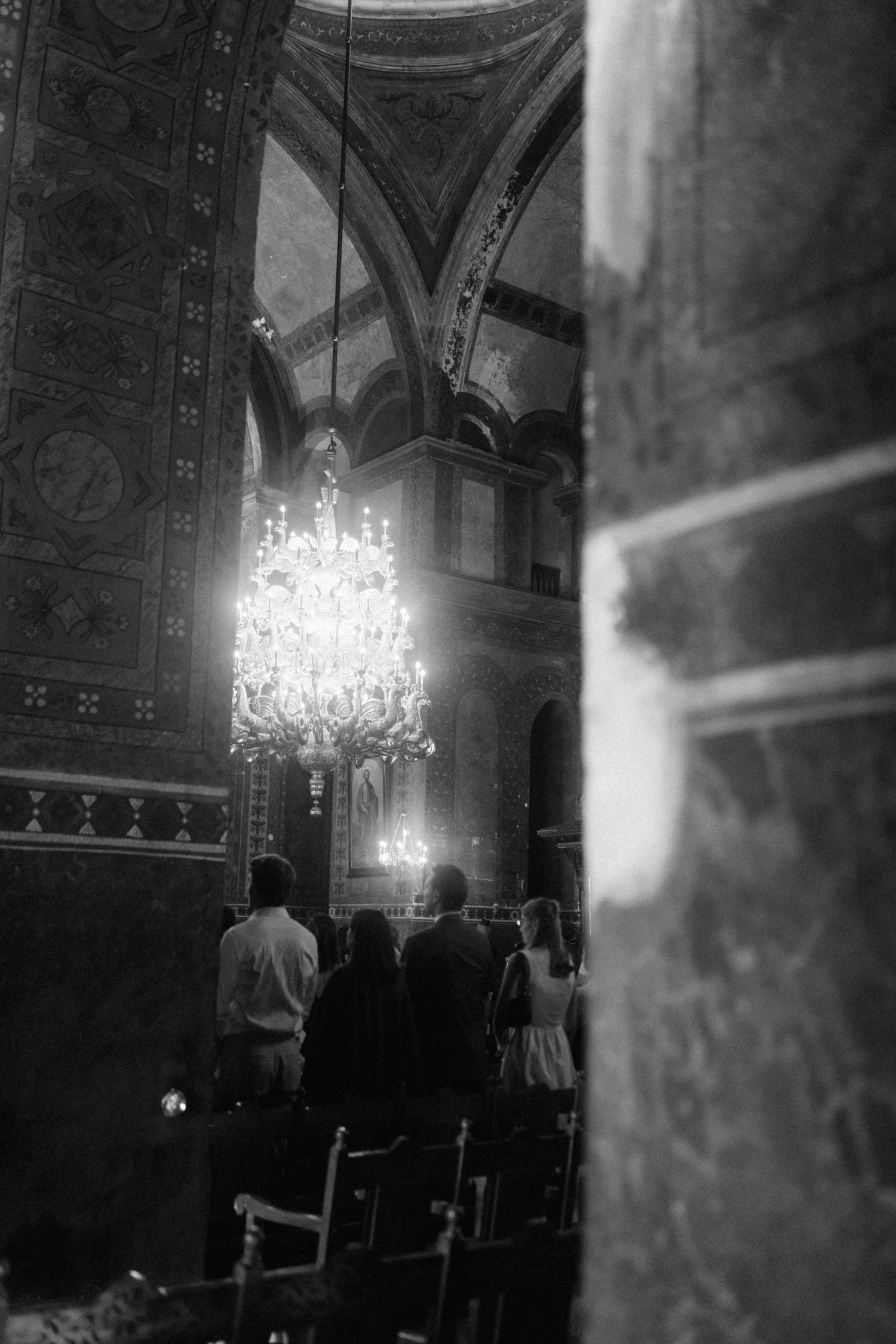 Black and white photo of wedding guests standing in ornate church with chandelier.