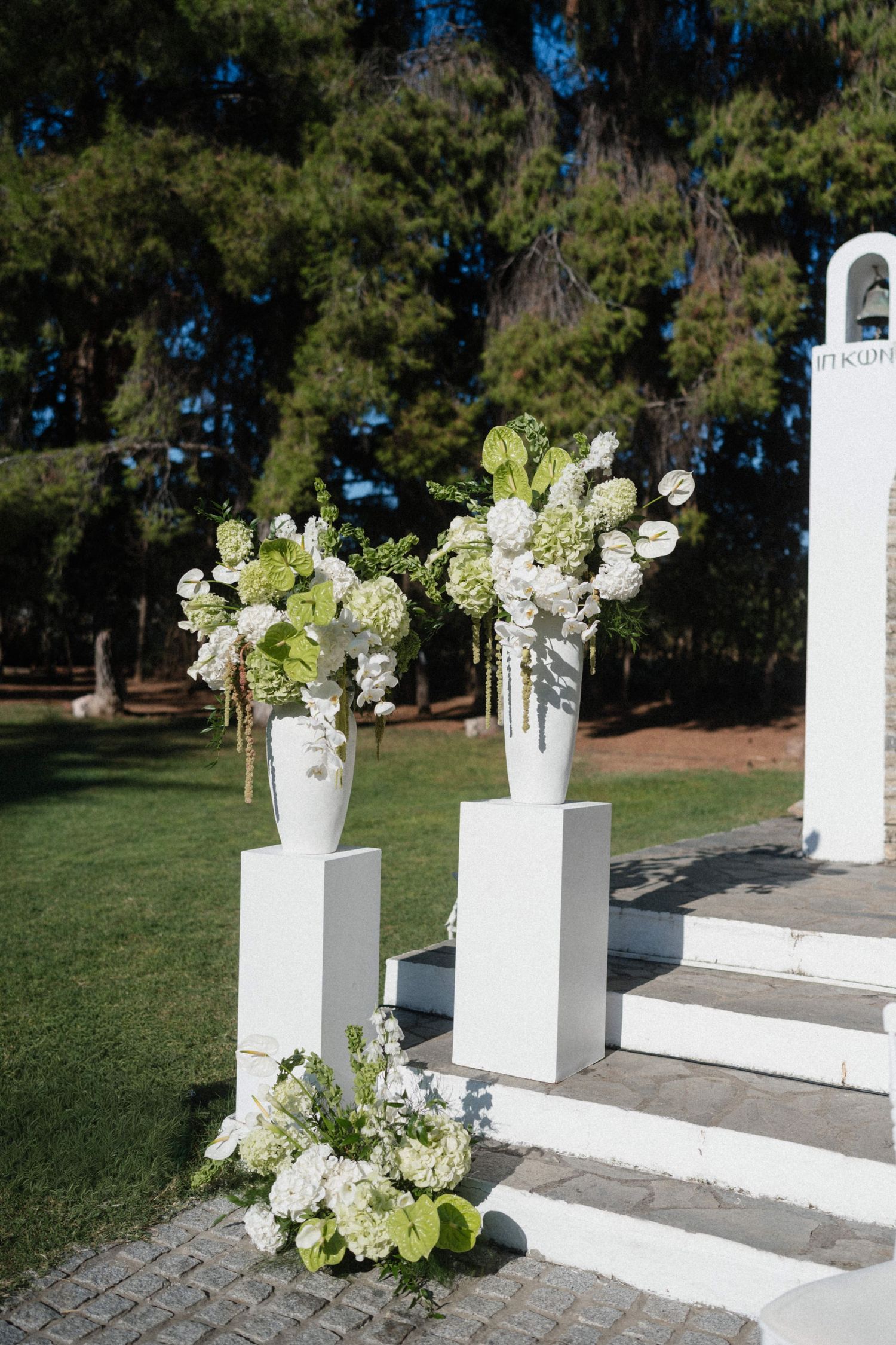 Two white vases with green and white floral arrangements on pedestals at an outdoor wedding ceremony.
