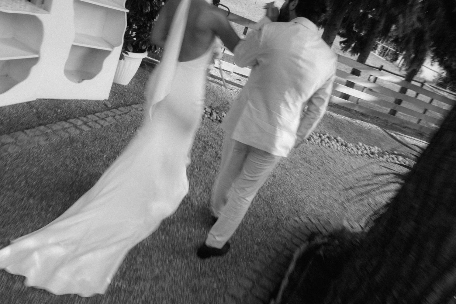 Black and white photo of a bride and groom walking outdoors on their wedding day, bride's train trailing behind.