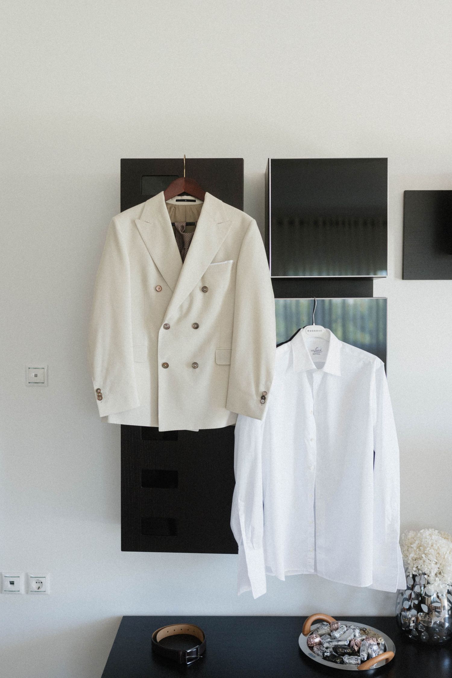 White double-breasted blazer and dress shirt hanging on wall hooks above a dark console table.