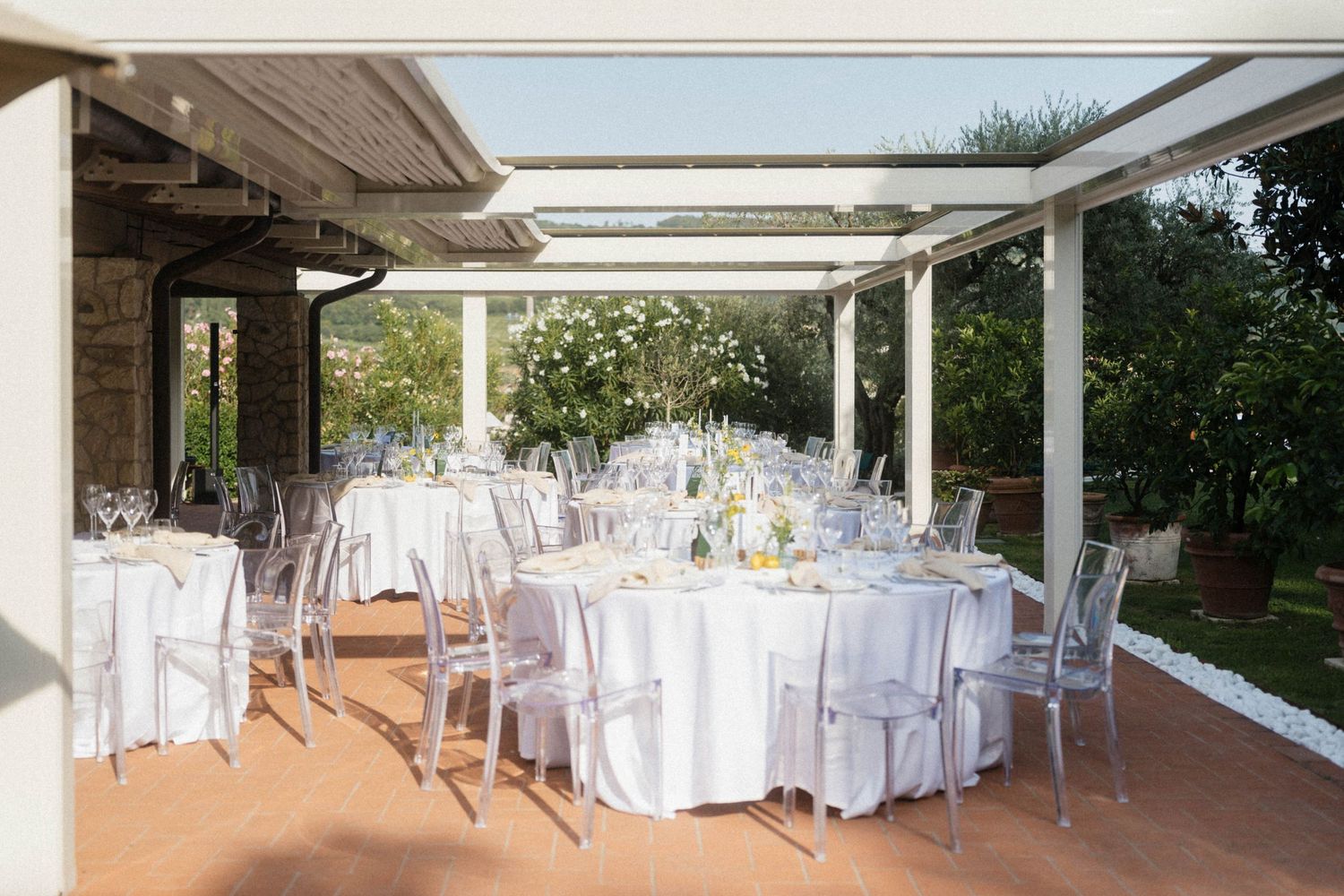 Elegant outdoor wedding reception with white round tables, ghost chairs, and floral centerpieces under a pergola.