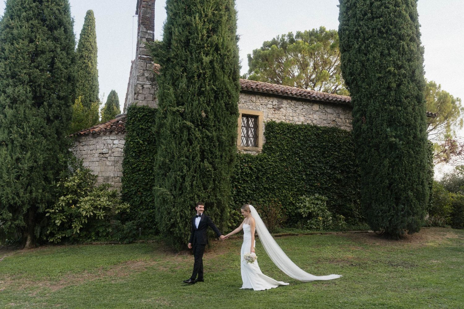 Bride and groom hold hands in front of a stone villa surrounded by tall cypress trees in Tuscany.