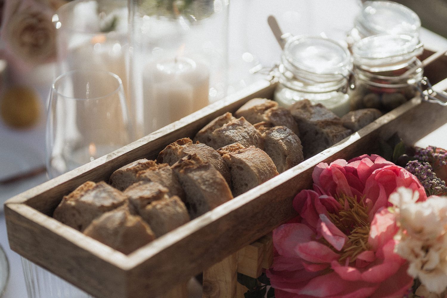 Rustic wooden tray filled with artisan bread rolls displayed on a table with candles and pink flowers.