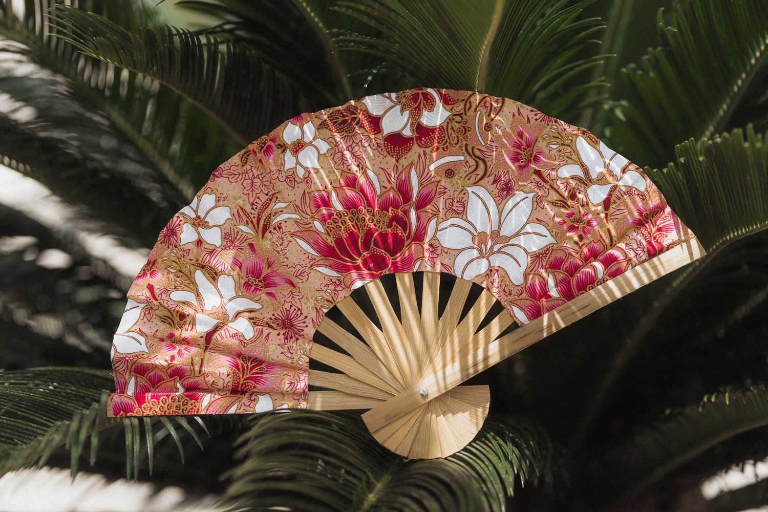 A decorative hand fan with pink and white floral patterns and gold accents, displayed against tropical green palm leaves.