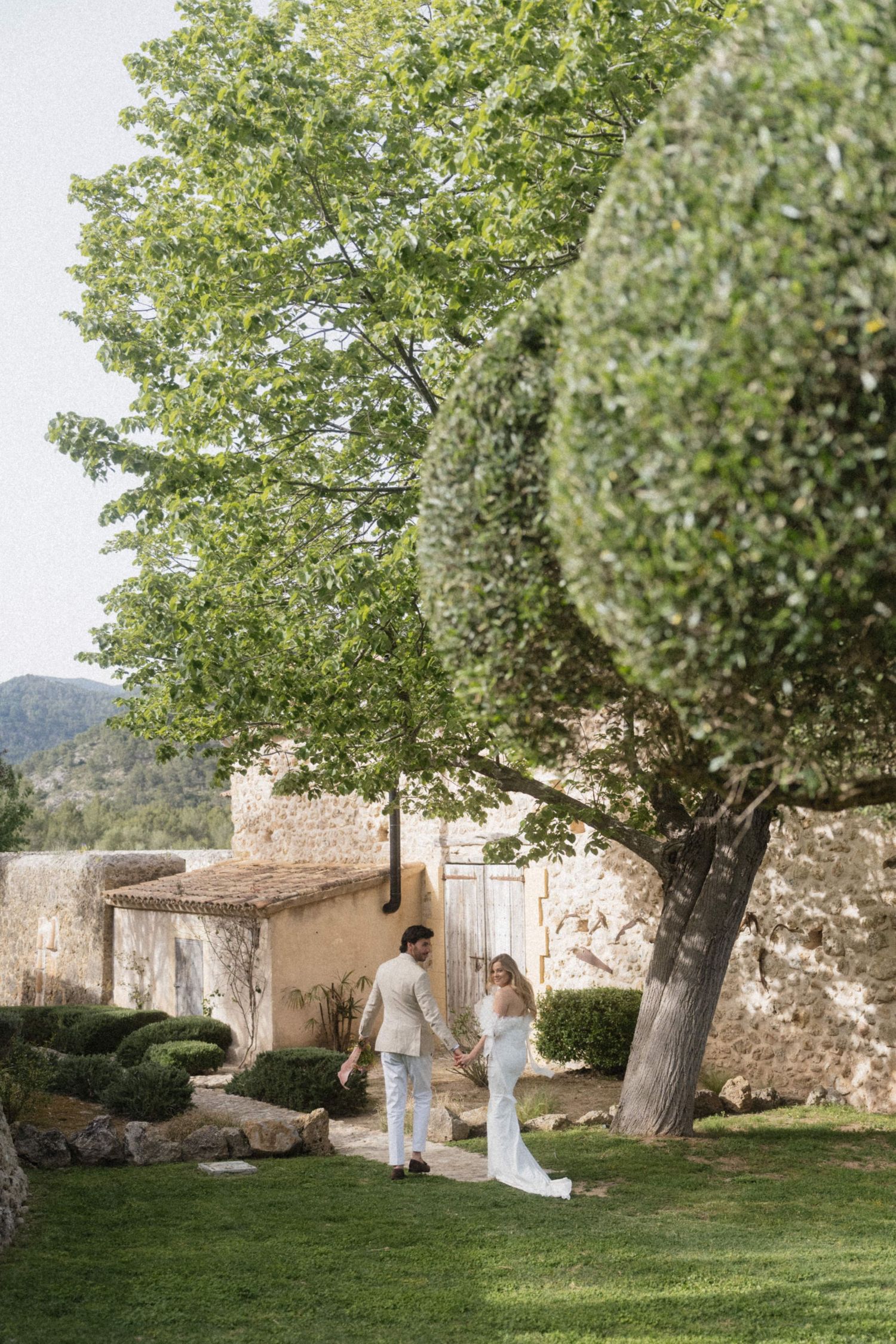 Bride and groom holding hands walking through a lush garden with stone walls and large trees at a Mallorca venue.